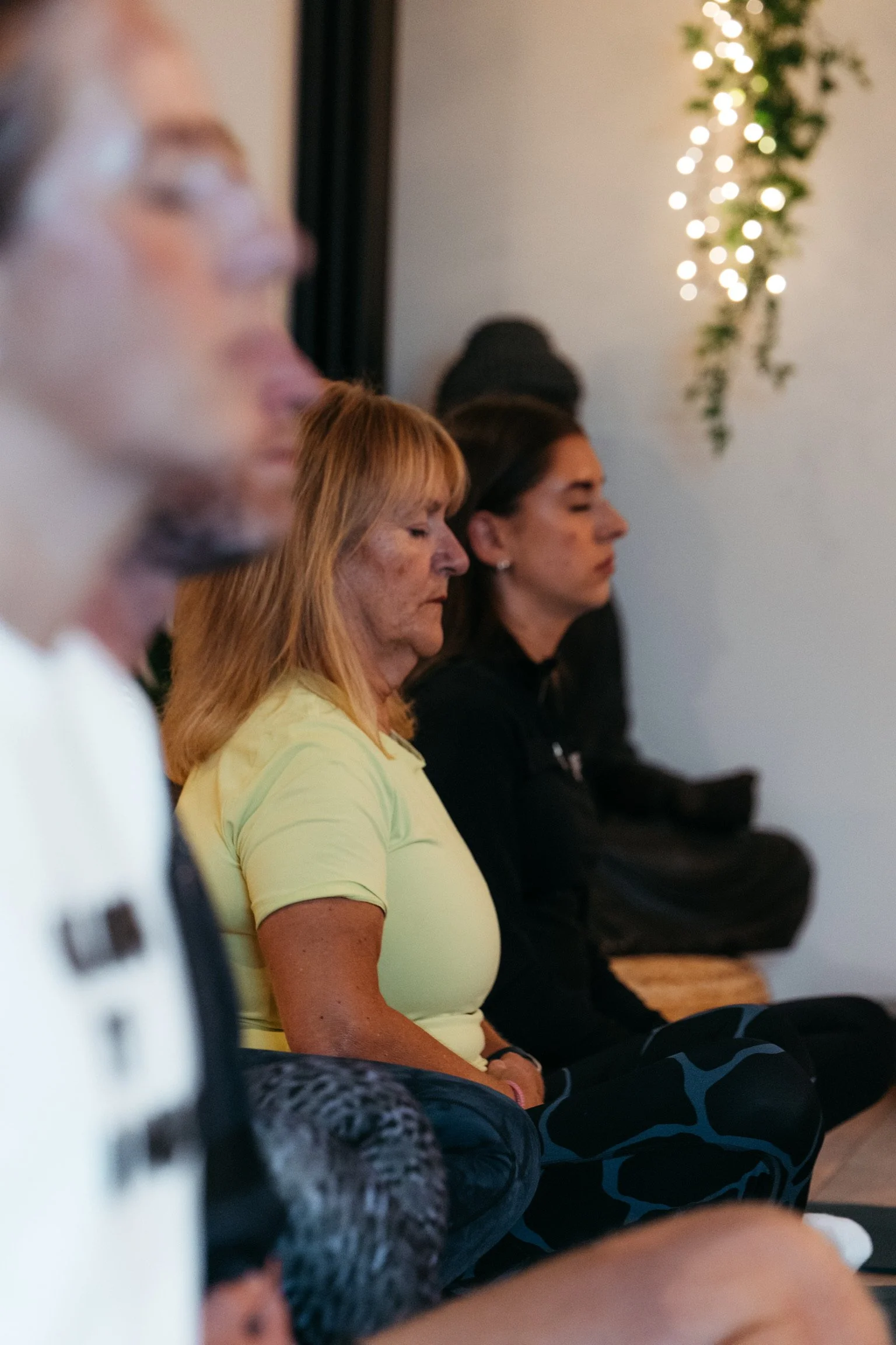 People sitting with eyes closed, possibly meditating or in prayer, inside a room with a wall decorated with greenery and string lights.