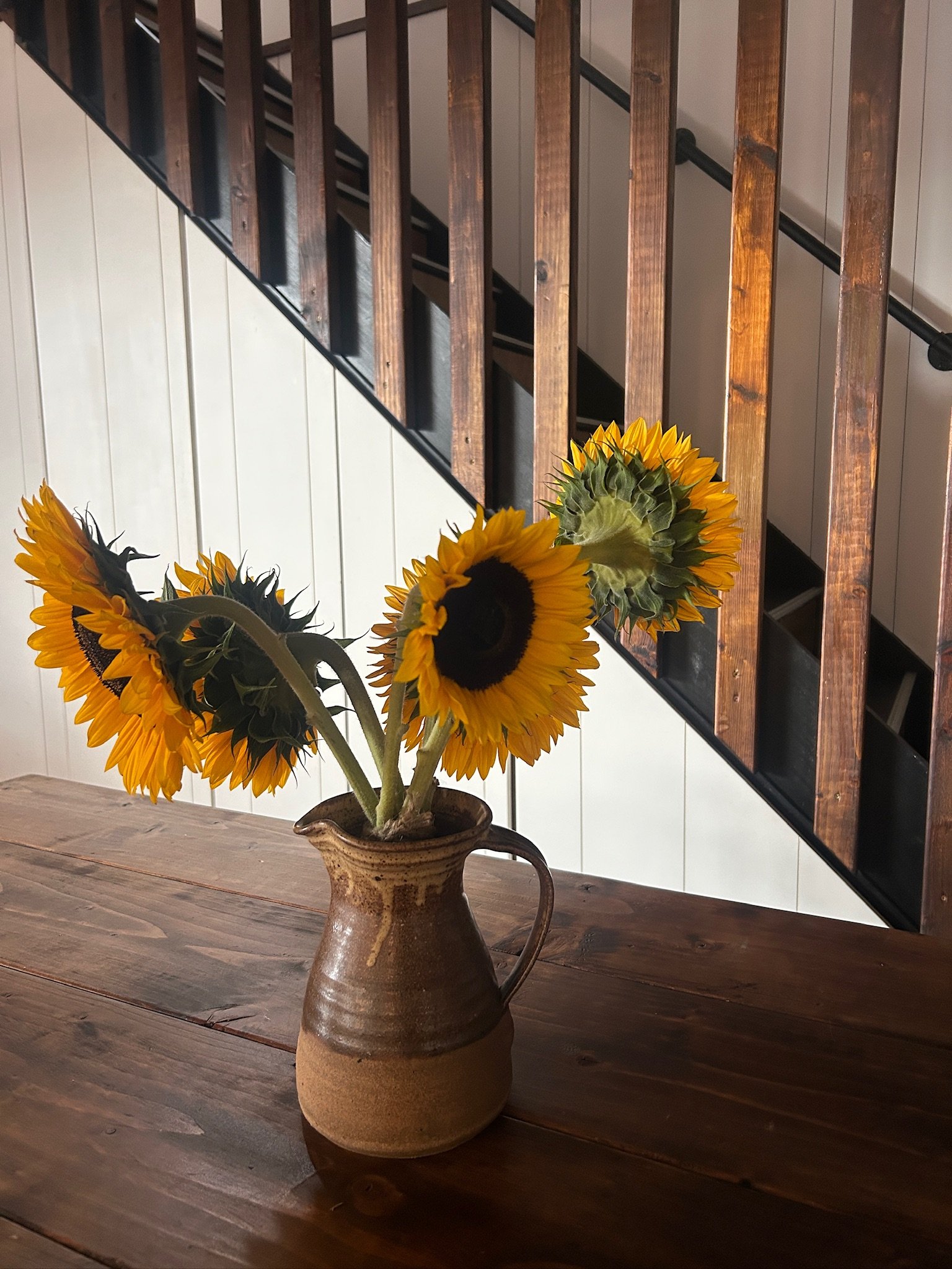 A ceramic vase with sunflowers on a wooden table, with a staircase and wooden railing in the background.