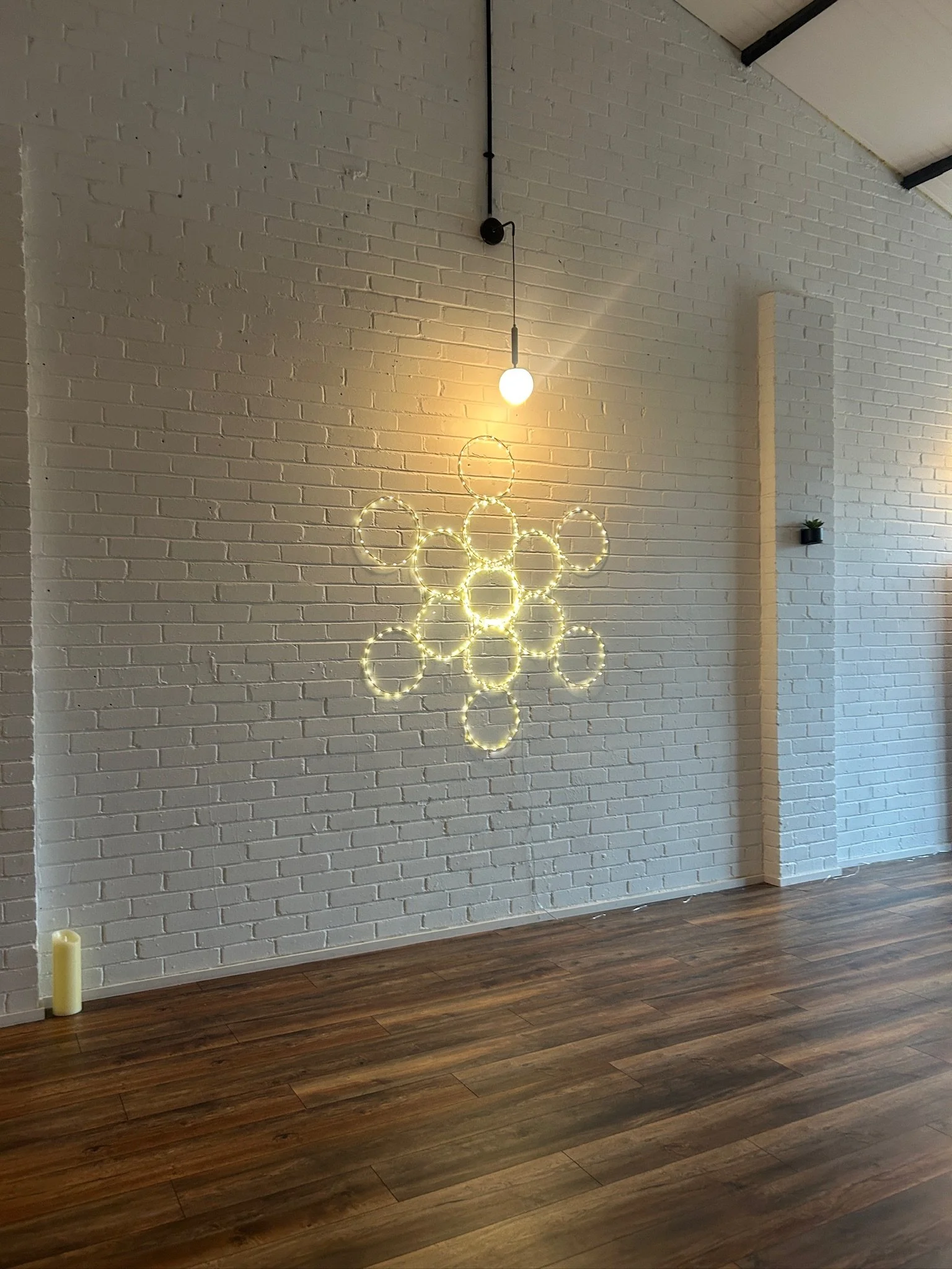 Decorative wall with a hanging LED light fixture shaped like a cluster of rings on a white brick wall in a room with wooden flooring.