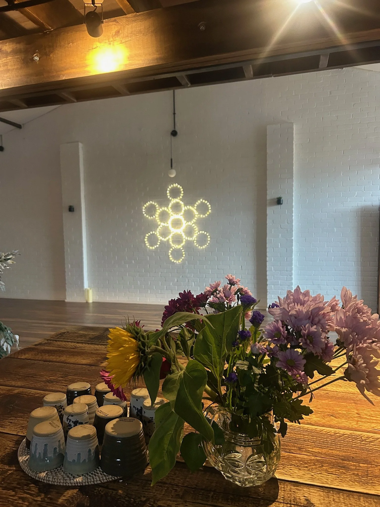 A rustic indoor space featuring a wooden table with a vase of colorful flowers and stacks of cups. In the background, a modern chandelier with circular lights hangs from a white brick wall.