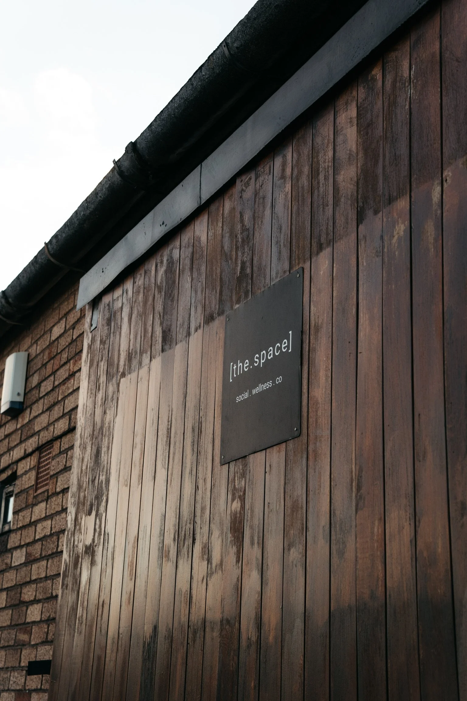 A wooden exterior of a building with a black sign that reads '[the.space] social.wellness.co' and a security camera on the left side.