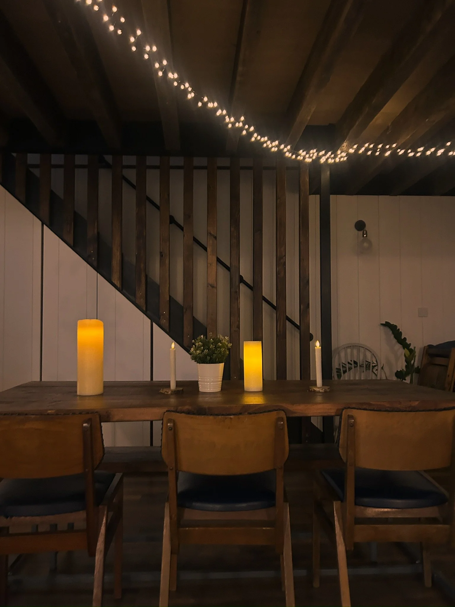 A cozy dining area with a rustic wooden table, four wooden chairs with dark cushions, and three candles and a potted plant as table decorations. There are string lights hanging from the ceiling and a staircase with wooden railings in the background.