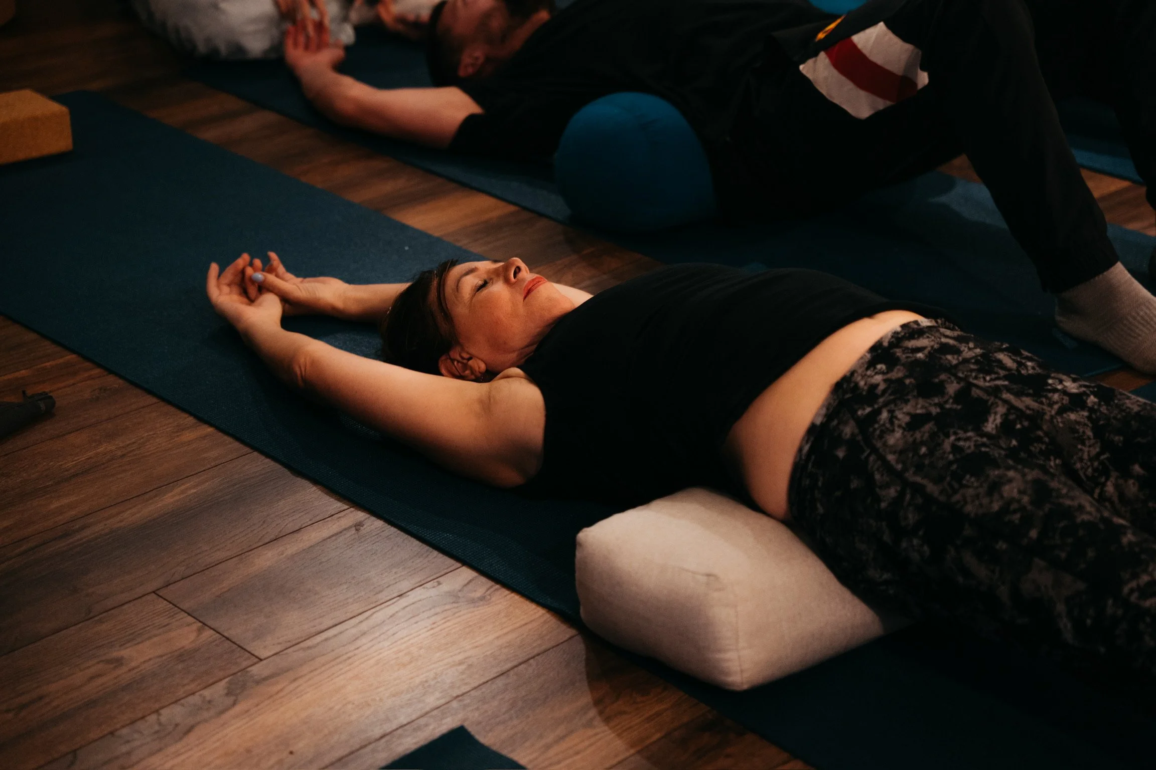 A woman lying on a yoga mat with eyes closed, arms stretched above her head, receiving a massage or therapy from a practitioner in a dark indoor setting.