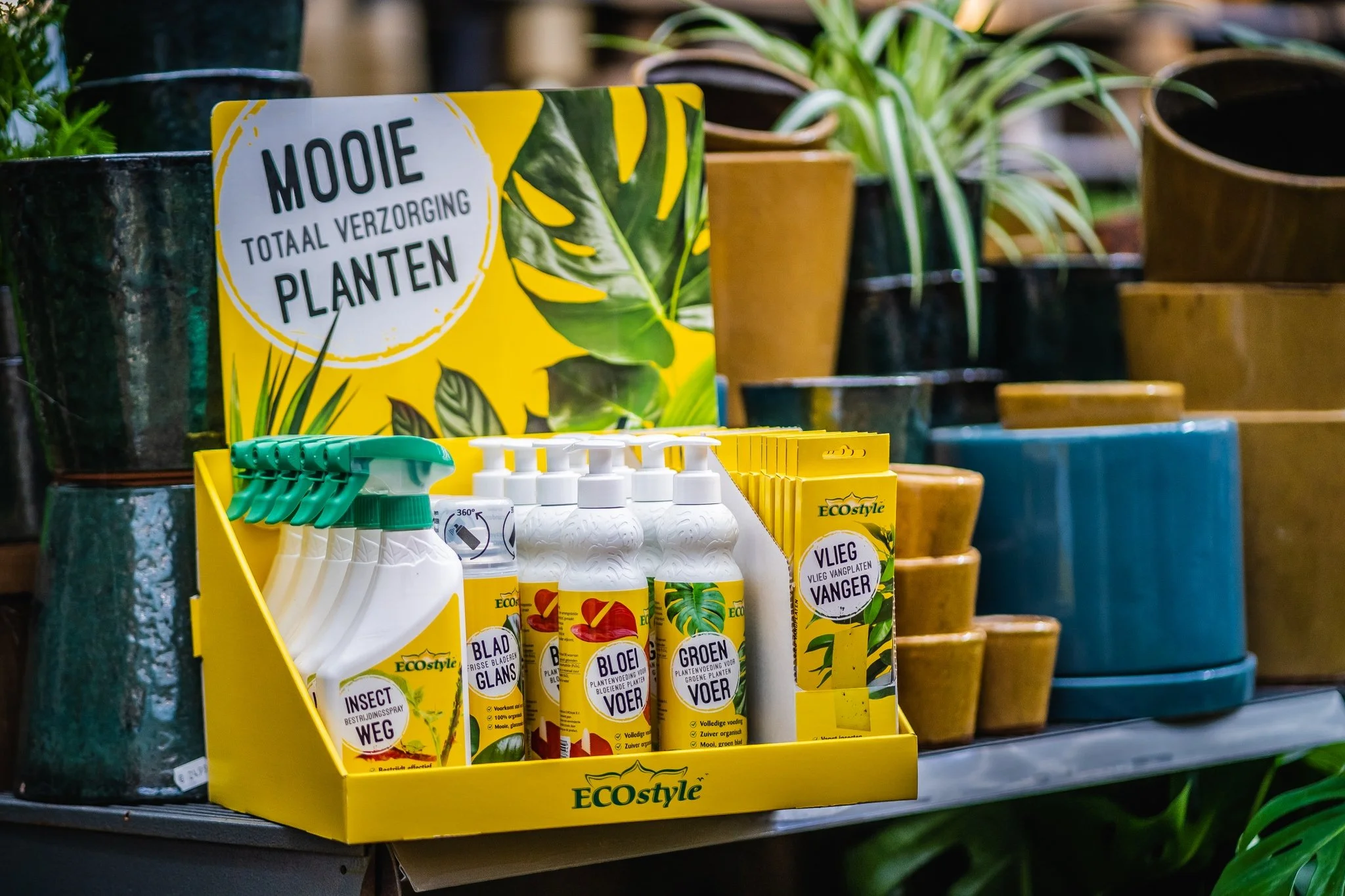 Verkoopdisplay met plantenverzorgingsproducten van ECOstyle, zoals plantensprays en -voeding, in een tuincentrum met verschillende potten en planten op de achtergrond.