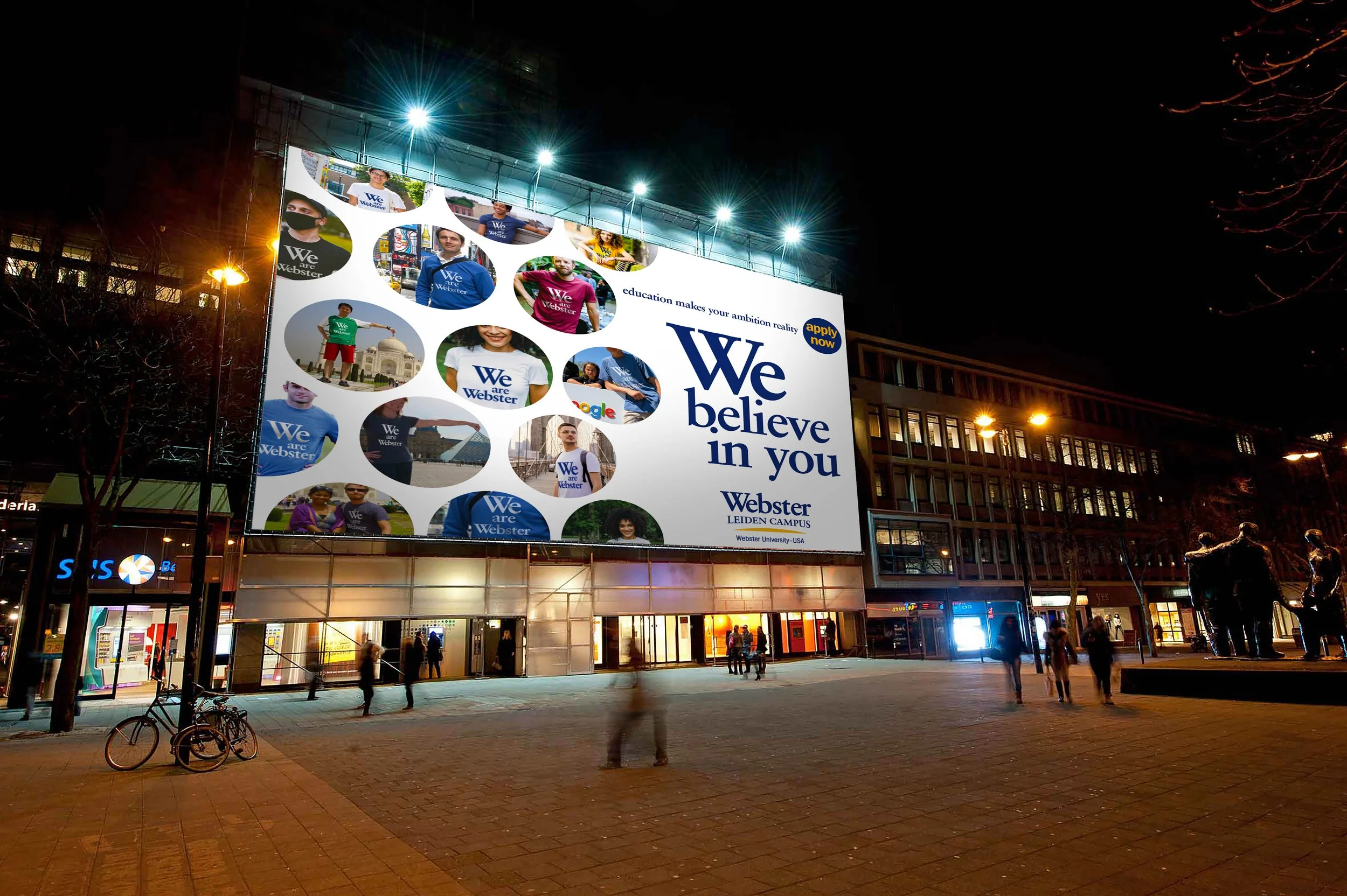 Een grote verlichte billboard op een stadsplein met mensen eromheen 's avonds. De billboard toont foto's van mensen die Webster University-merchandise dragen en de tekst 'We believe in you'.