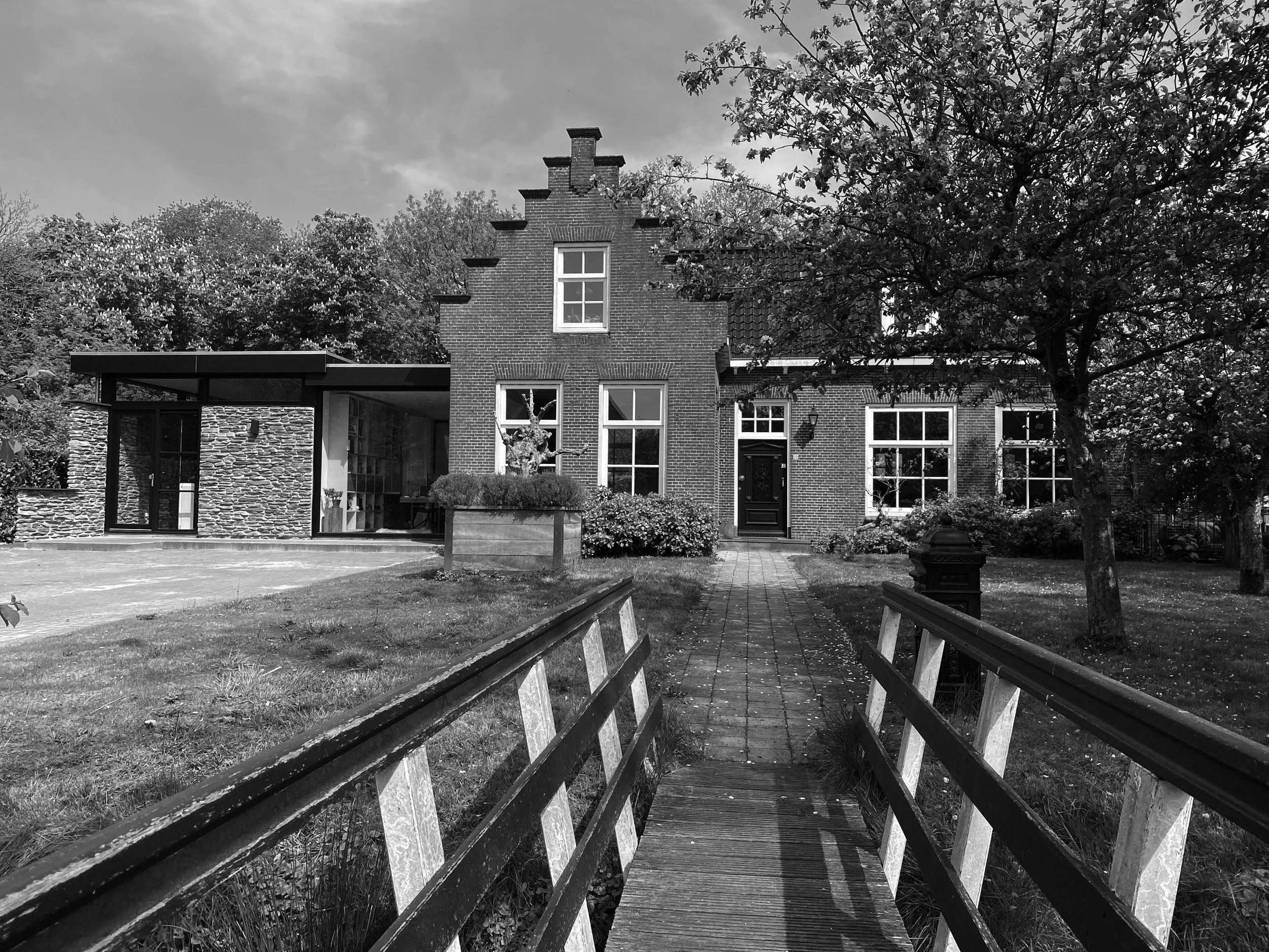 Een zwart-wit foto van een huis met een trap en bruggetje voor de ingang, omringd door bomen en een tuin.