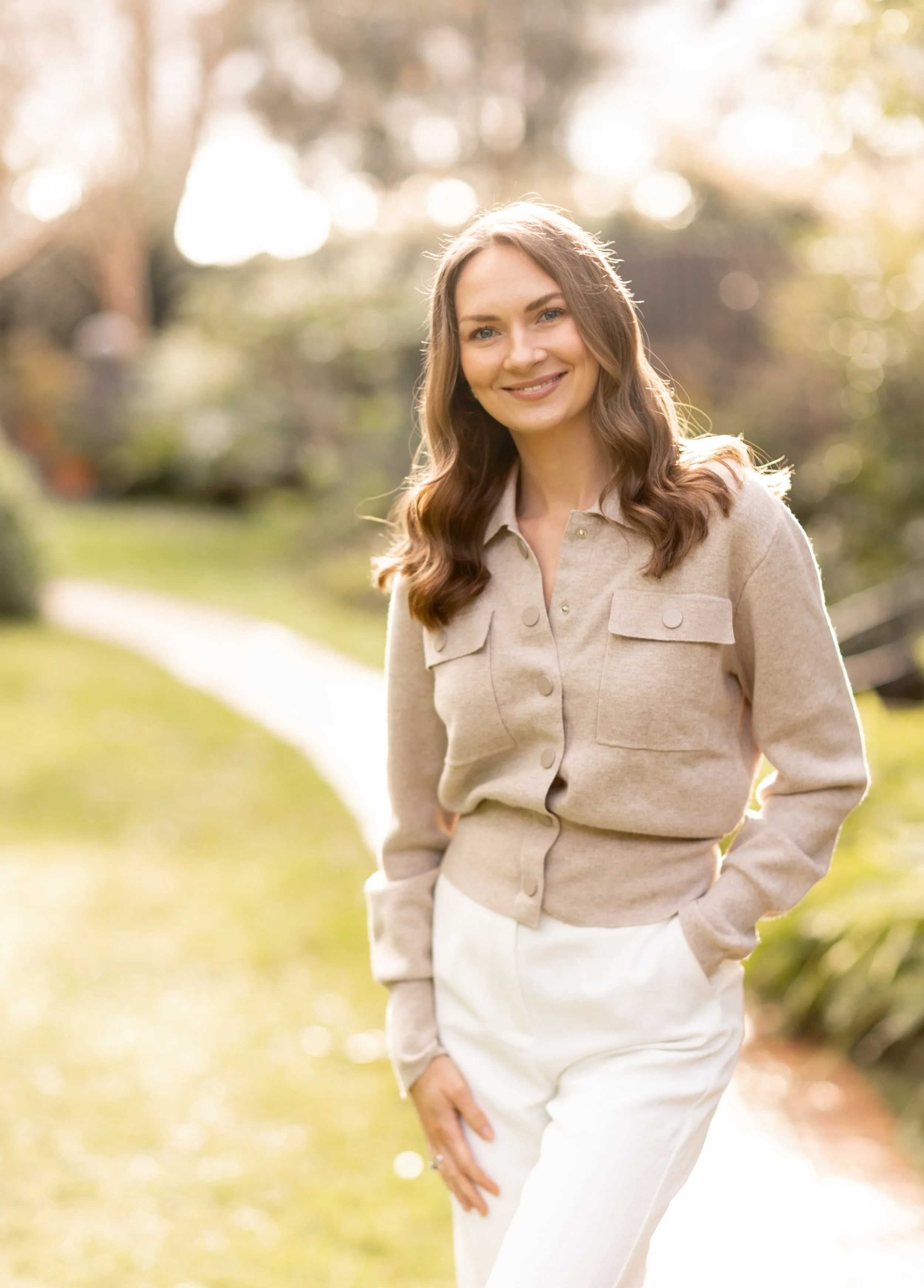 A woman with long, wavy brown hair smiling outdoors in a beige jacket and white pants on a sunny day.