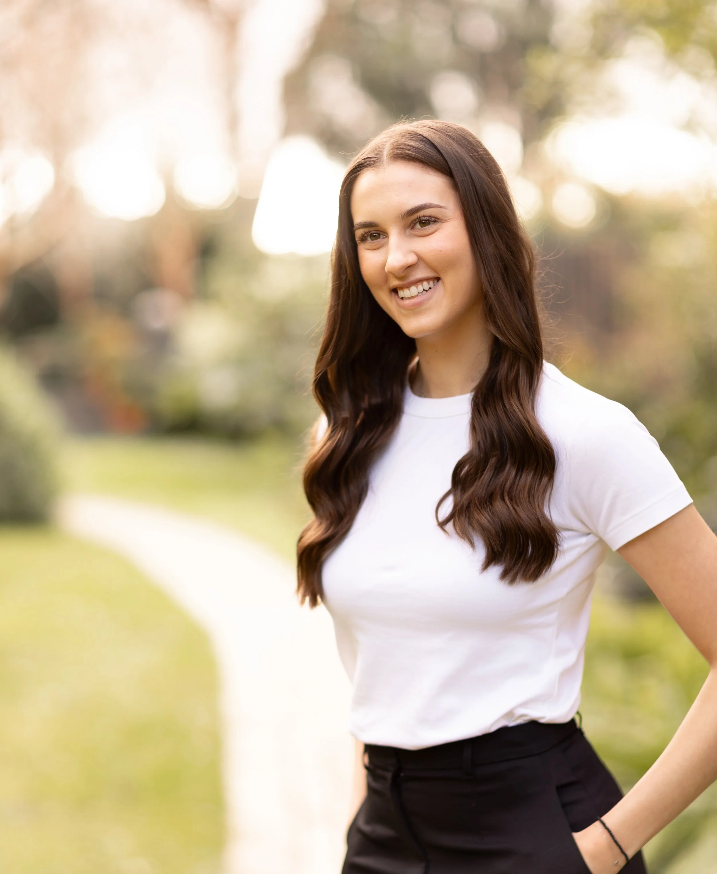 A young woman with long brown hair smiling outdoors in a park, wearing a white t-shirt and black pants.