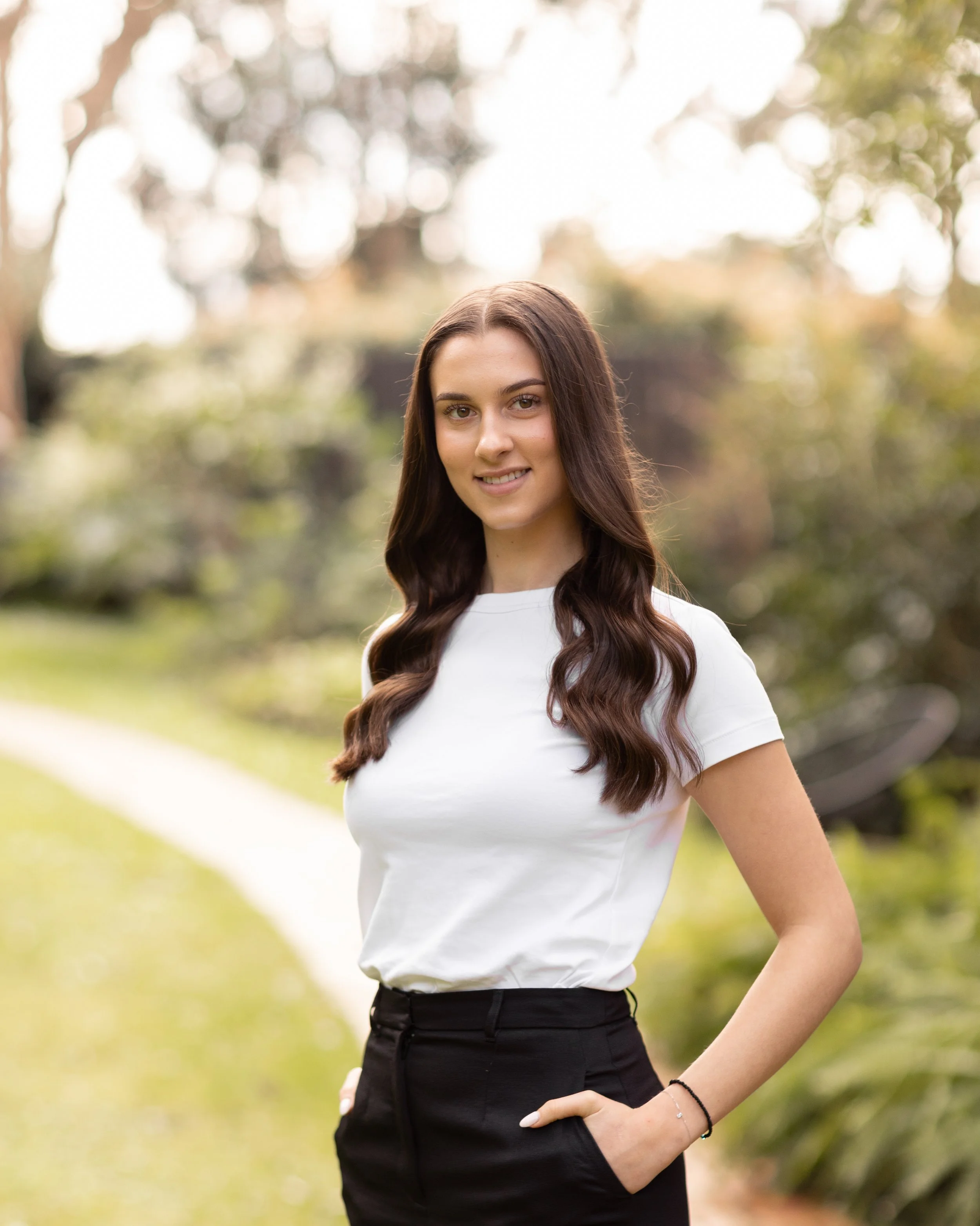 A young woman with long wavy brown hair, wearing a white t-shirt and black pants, standing outdoors on a trail in a park with trees and greenery in the background, smiling at the camera.