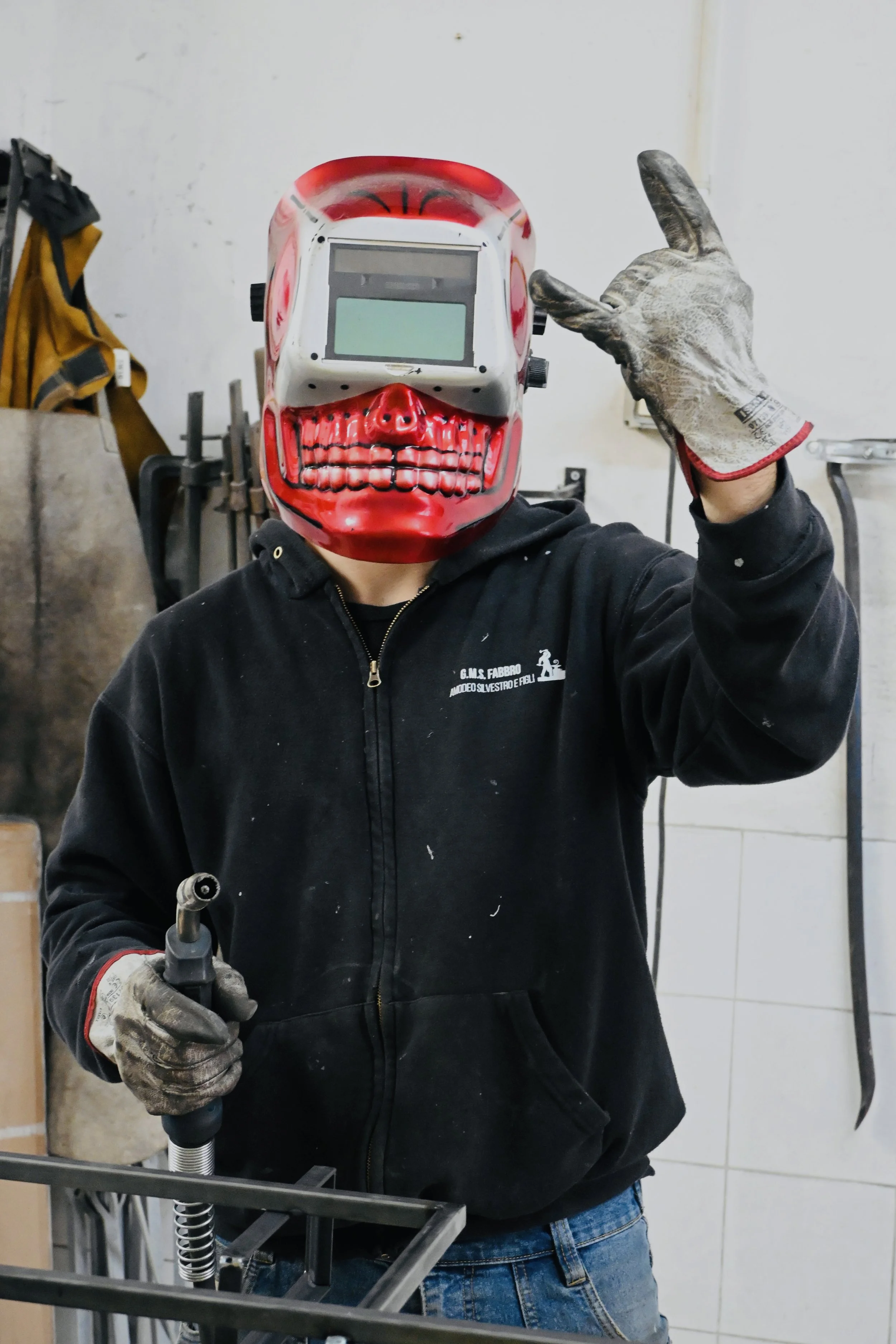 Persona con maschera da saldatore a forma di teschio, che indossa guanti da lavoro, tiene una pinza da saldatore e lavora in un'officina.