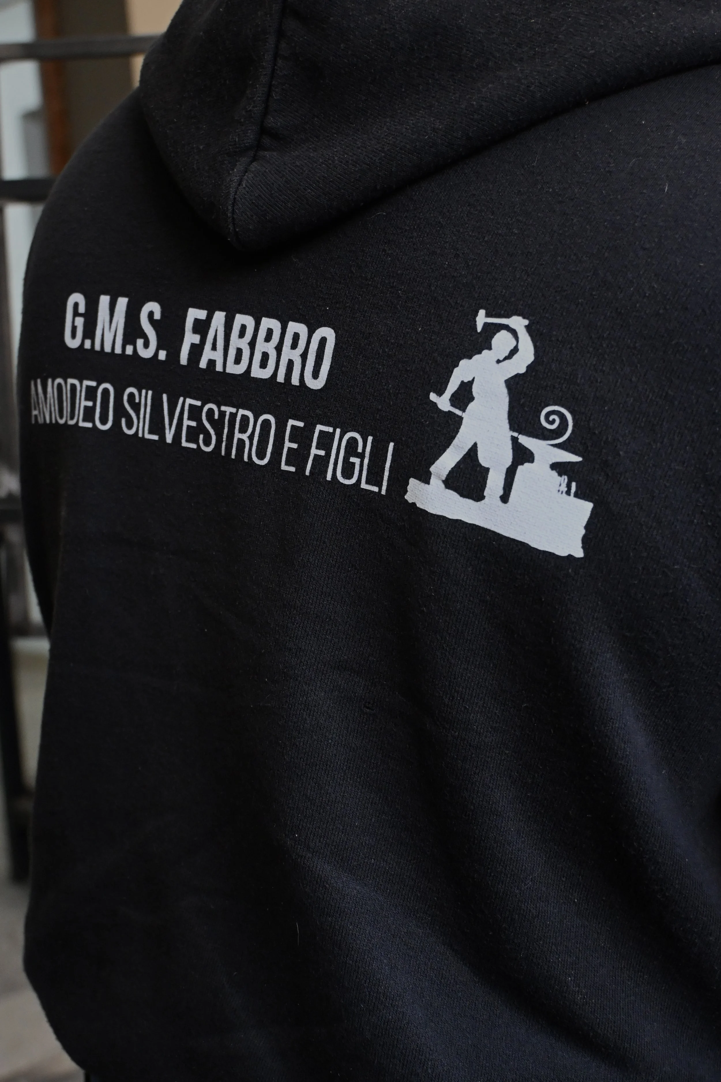 Zoom su una felpa nera con scritte bianche e un disegno di un fabbro che lavora, appartenente a un'associazione chiamata G.M.S. Fabbro Amodeo Silvestro e Figli.