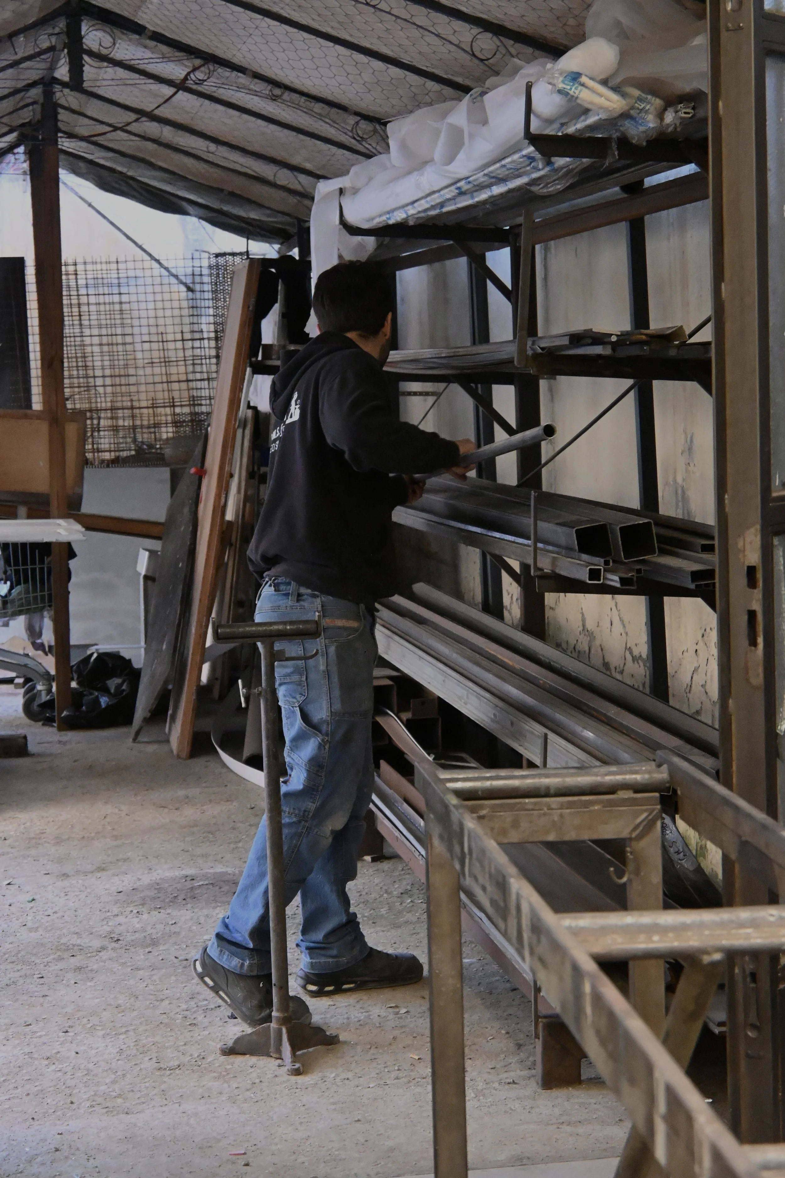 Un uomo indossa una felpa nera e jeans e lavora in un'officina o magazzino con ripiani di metallo pieni di tubi e materiali vari