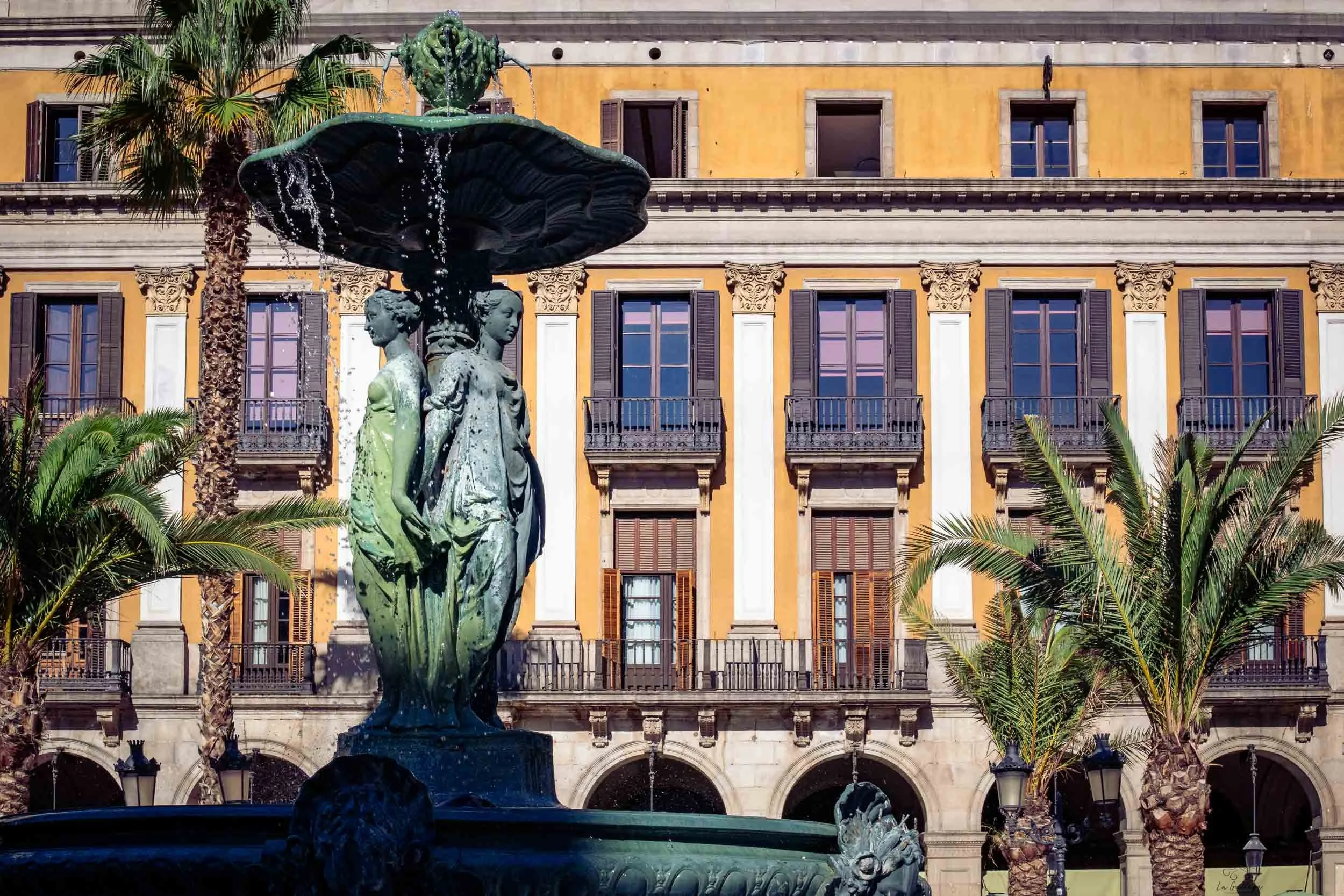 Decorative fountain with three female statues in Plaça Reial, Barcelona, framed by palm trees and a historic building.