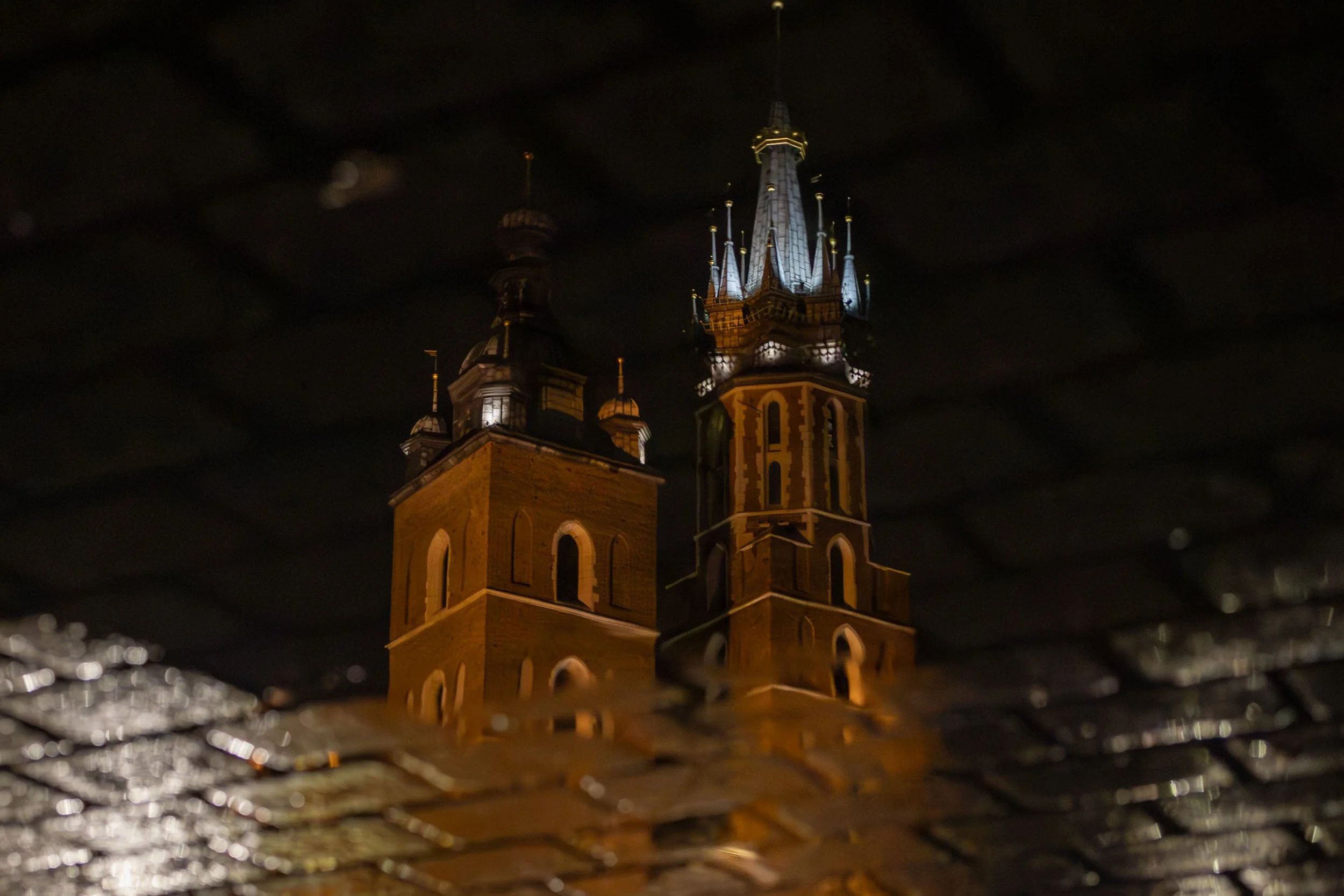 Towers of St. Mary’s Basilica in Krakow seen at night through a wet surface, with warm lights reflected in rain-soaked stone in the foreground.