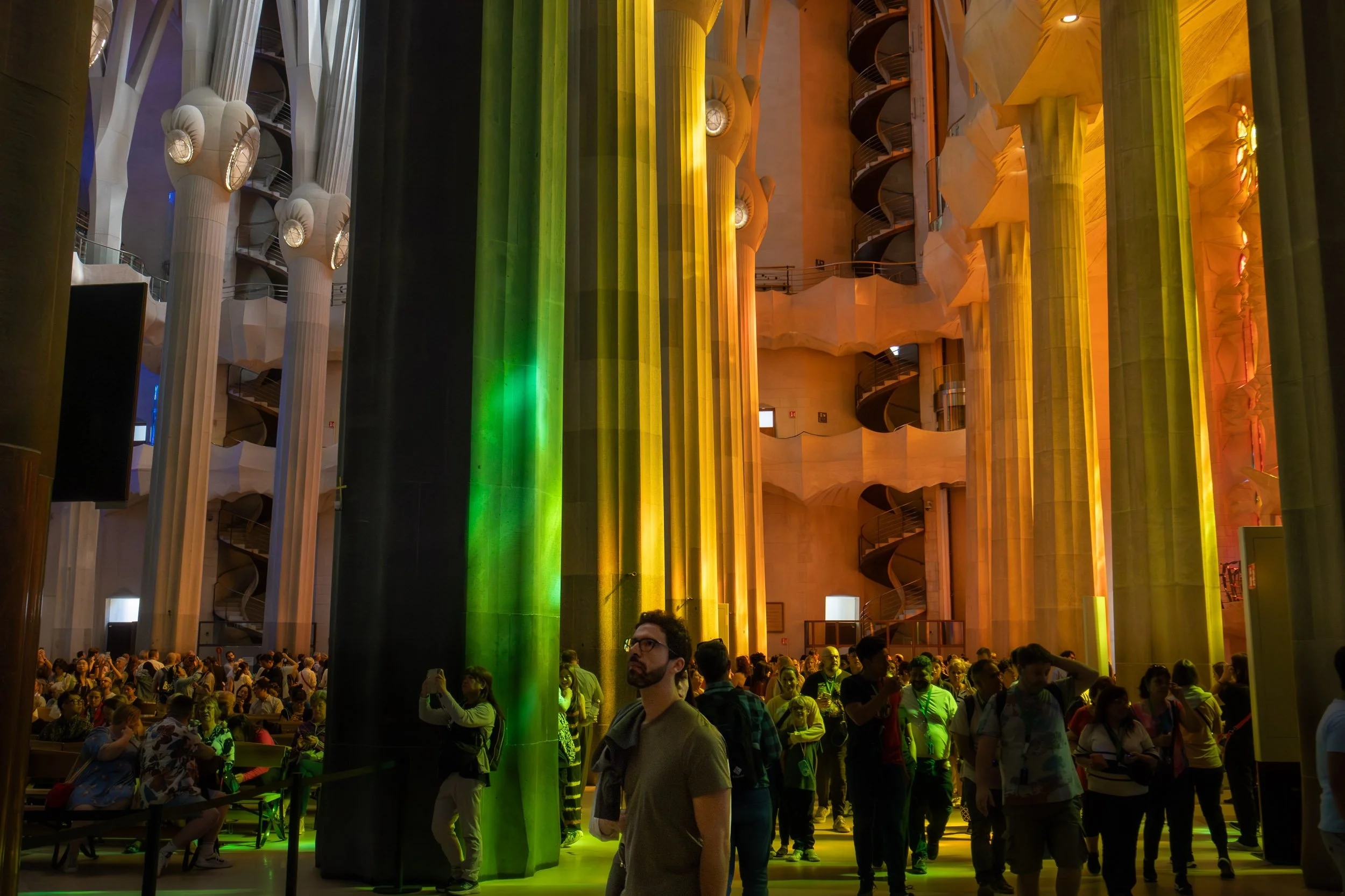 Interior view of La Sagrada Família in Barcelona, showing visitors walking among tall stone columns illuminated by green, yellow, and orange light filtering through stained glass windows.