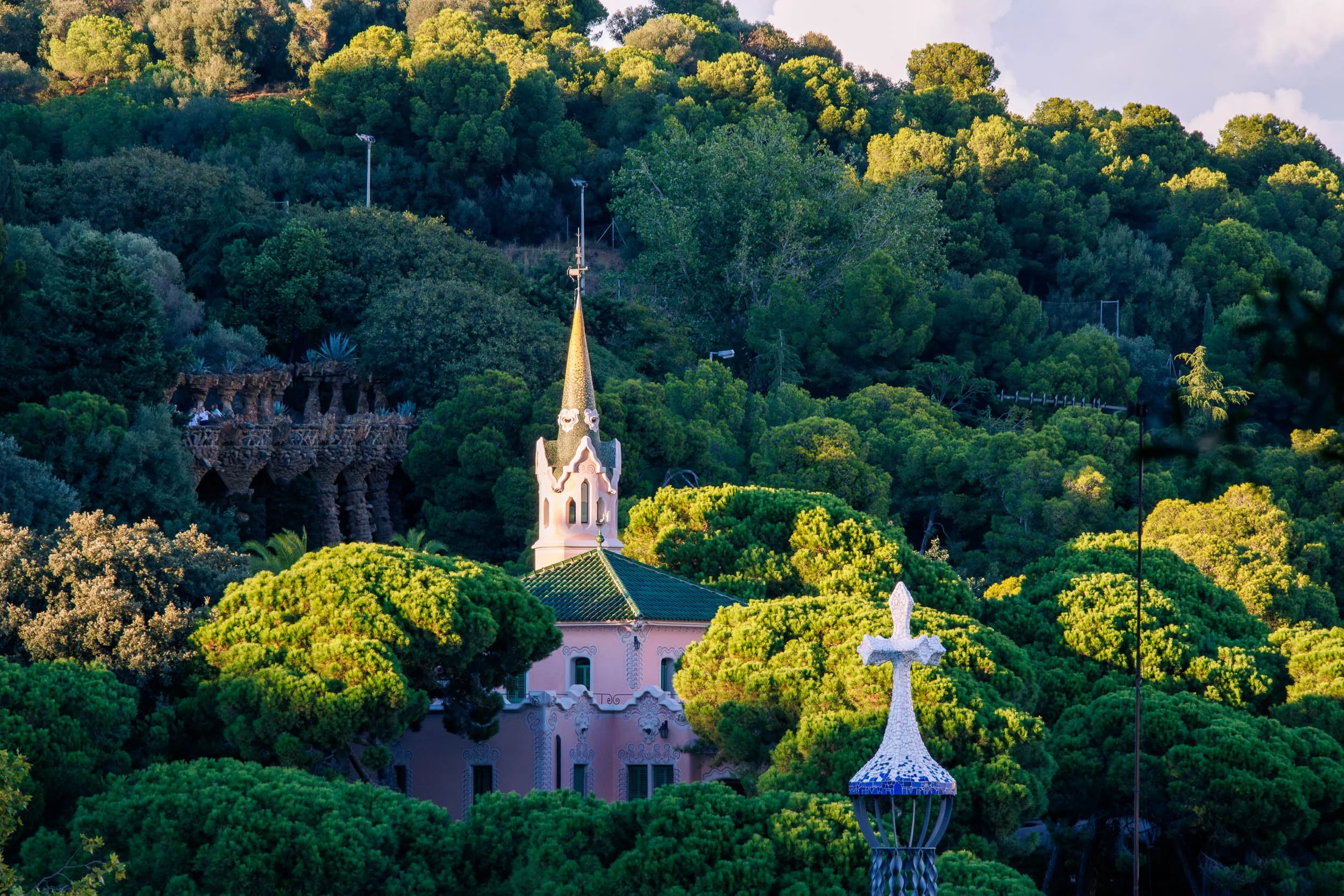 View of distinctive architecture in Parc Güell, showing a pastel-colored building with a slender tower and mosaic cross nestled among dense green trees.