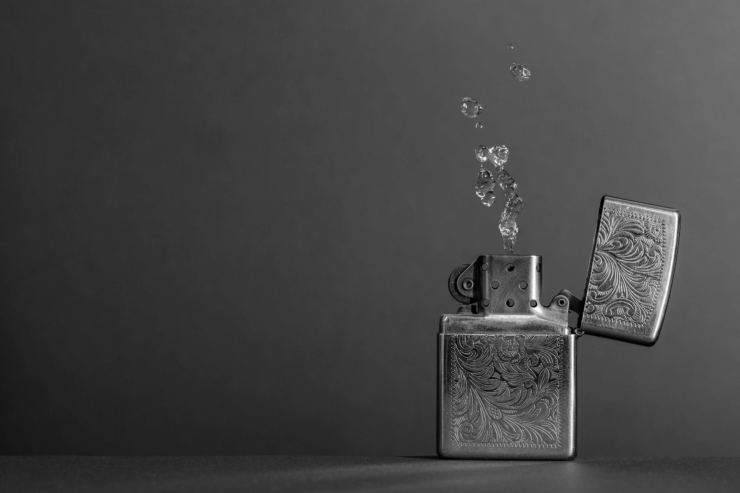 Conceptual black-and-white photograph of an open metal lighter producing a stream of water droplets instead of a flame, creating a surreal visual contrast.