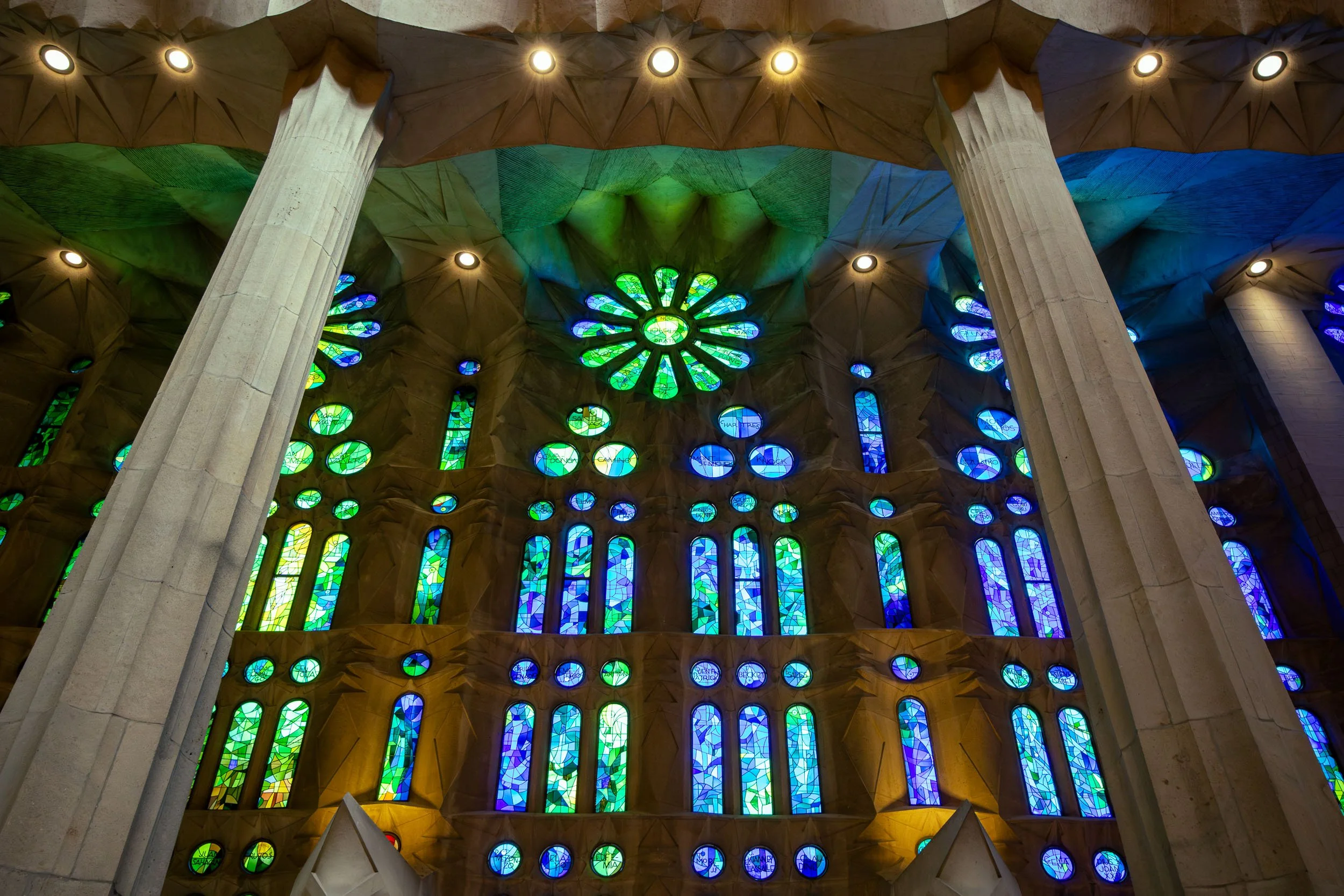 Interior view of La Sagrada Família in Barcelona, showing tall stone columns and vibrant stained glass windows casting blue and green light across the nave.