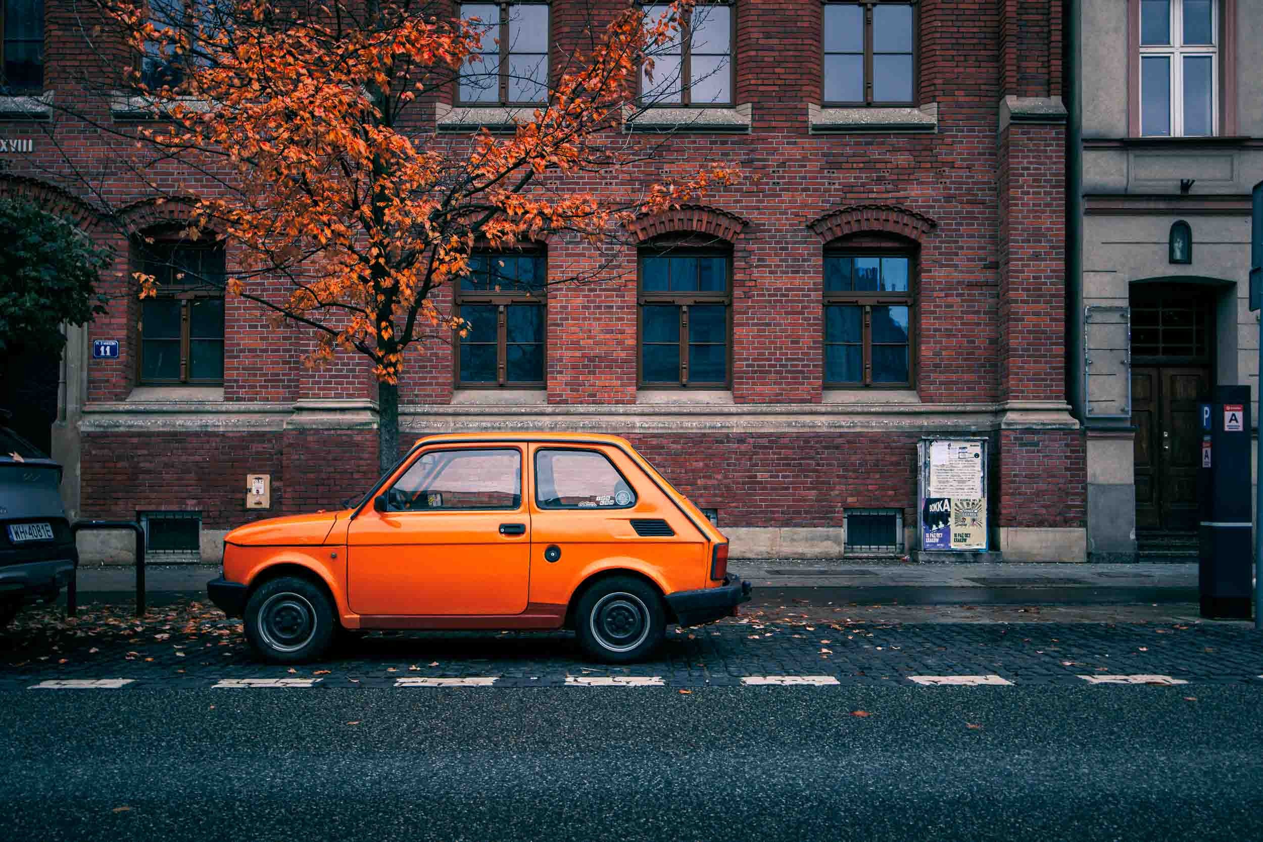 Small orange car parked on a cobblestone street in Krakow, framed by a brick building and an autumn tree with orange leaves.