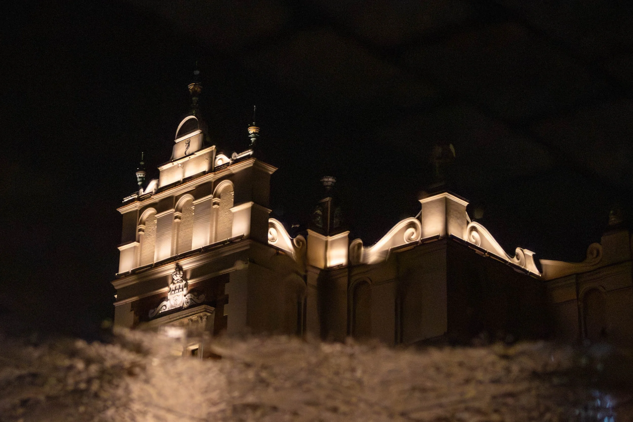 Illuminated facade of the Kraków Cloth Hall seen at night through a wet surface, with warm architectural lighting contrasting against the dark sky.
