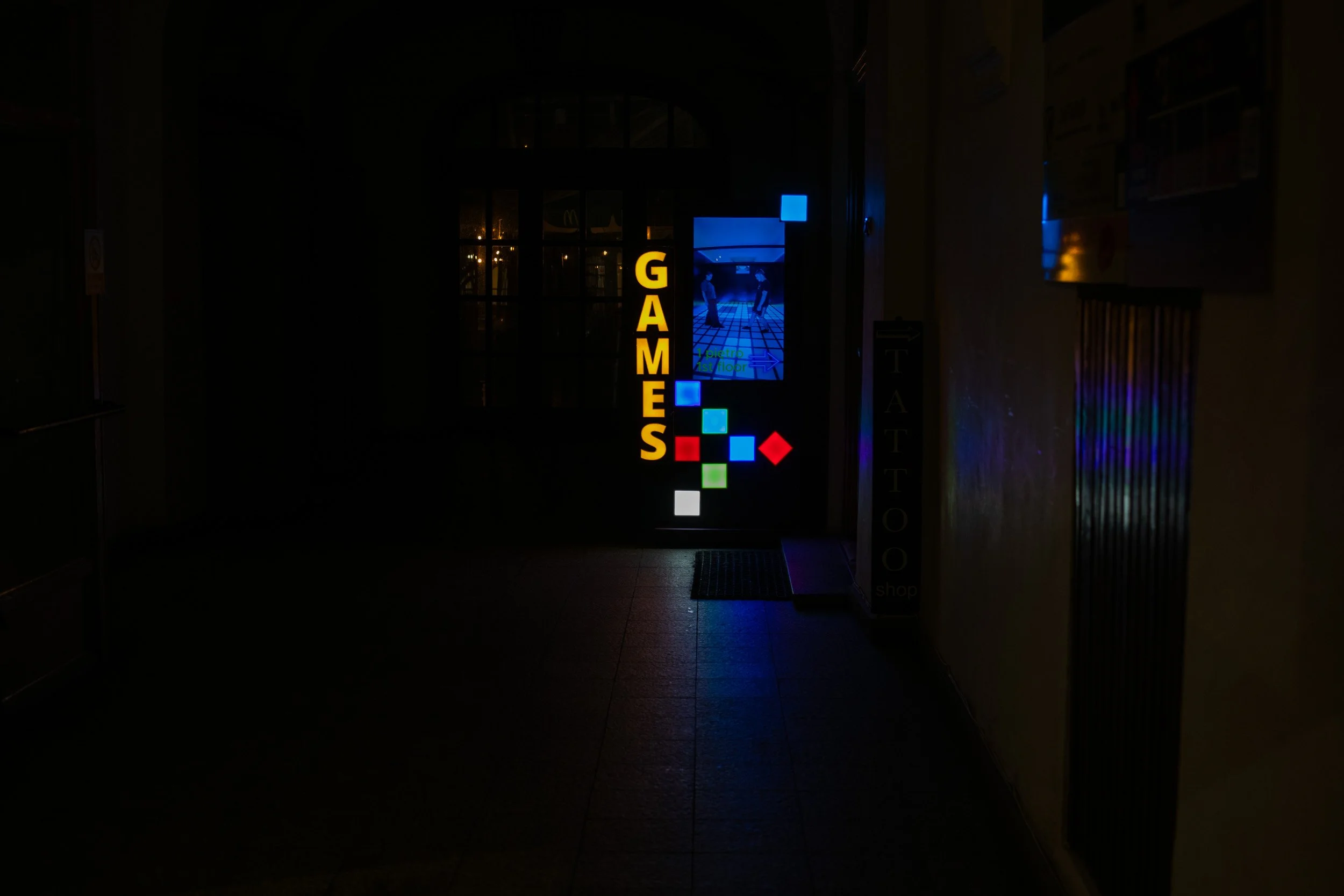 Illuminated “Games” sign with colorful geometric lights in a narrow passage at night in Krakow, surrounded by dark walls and reflected on the pavement.