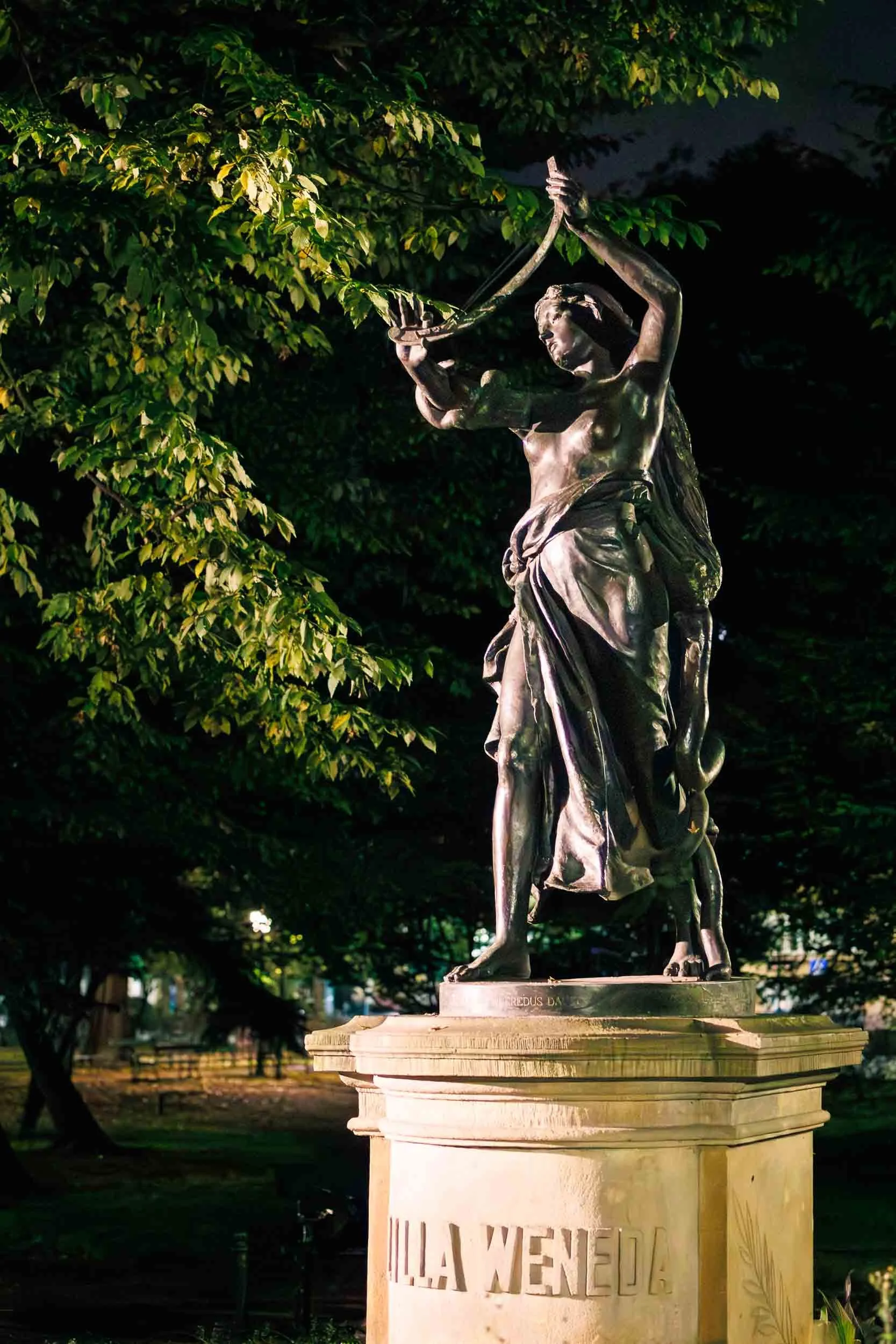 Bronze statue of a woman holding a harp, illuminated at night on a stone pedestal in a park in Krakow, surrounded by trees and dark foliage.