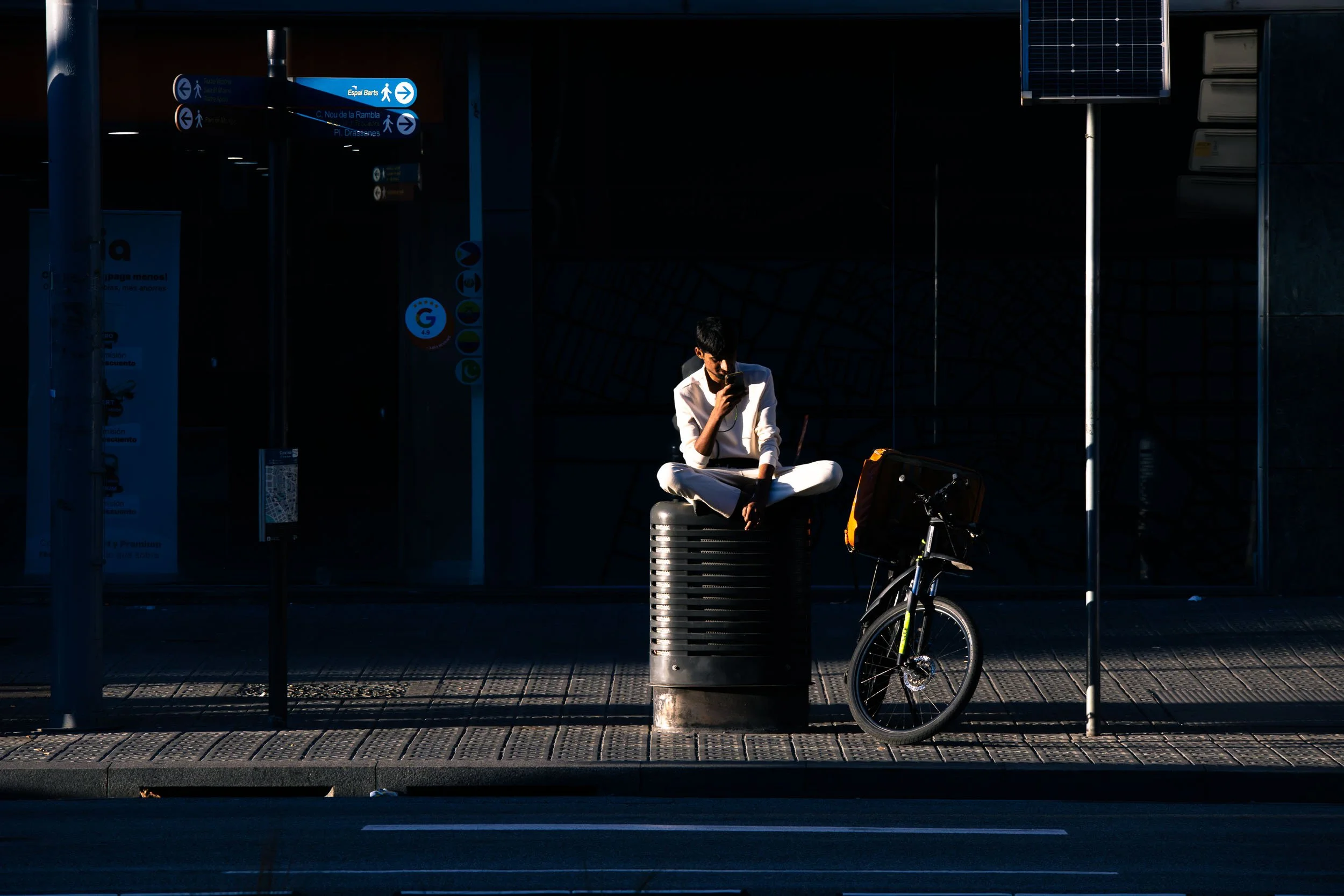 Person sitting on a trash bin beside a bicycle in the Poble-sec neighborhood of Barcelona.