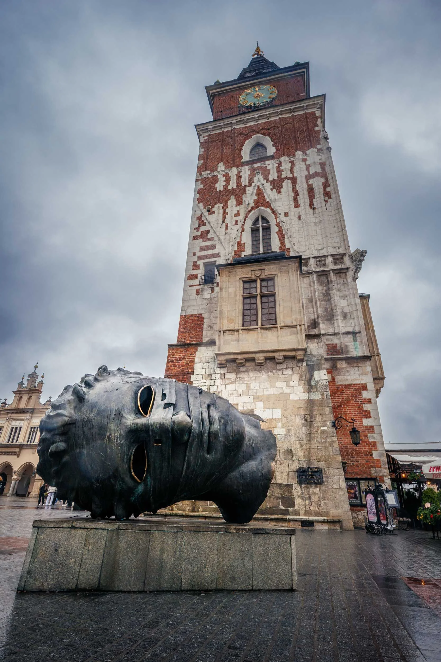 Large bronze head sculpture lying on a stone pedestal in front of the Town Hall Tower on a city square in Krakow under an overcast sky.