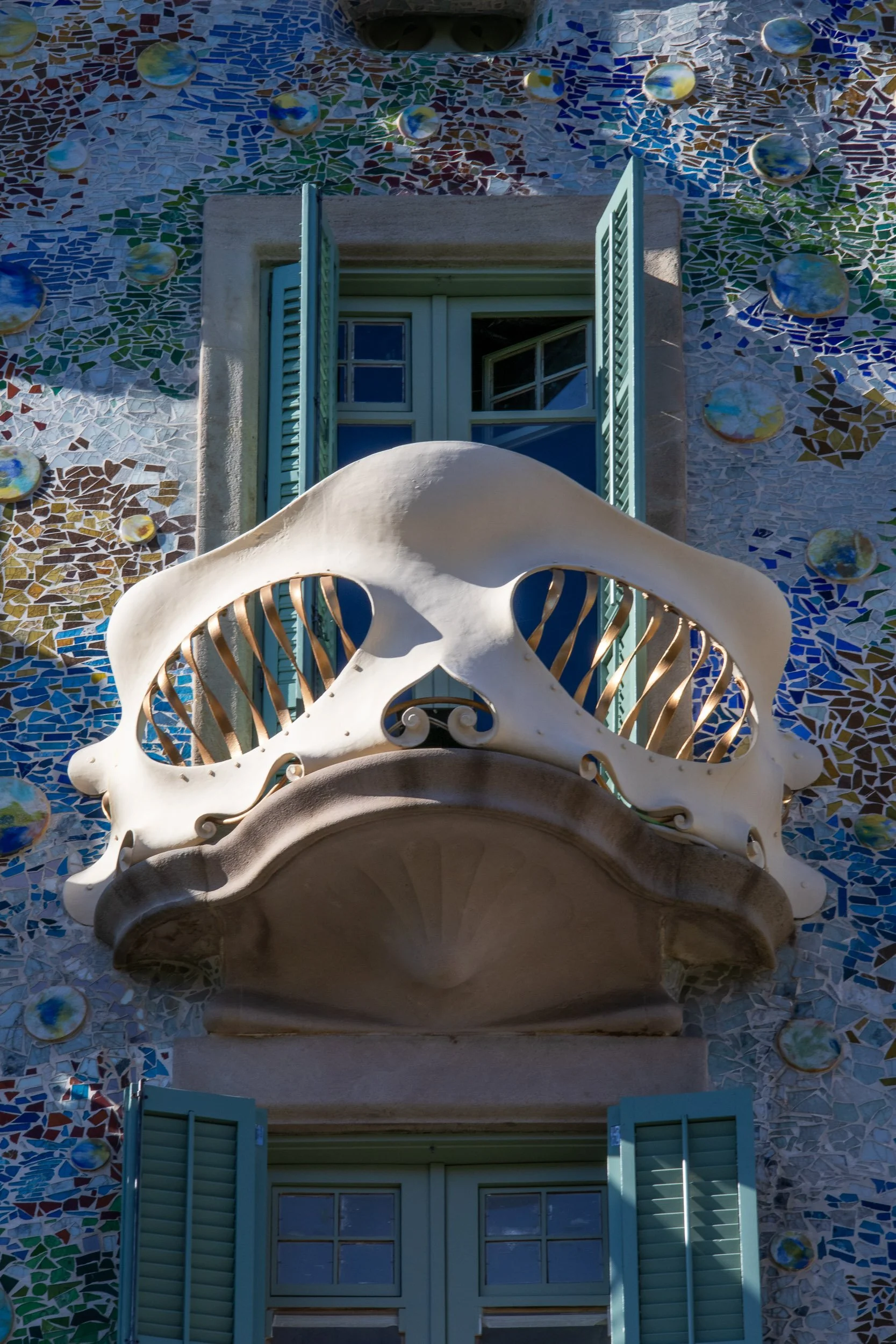 Close-up of a sculptural balcony on the facade of Casa Batlló in Barcelona, with organic shapes, colorful mosaic tiles, and shuttered windows behind.