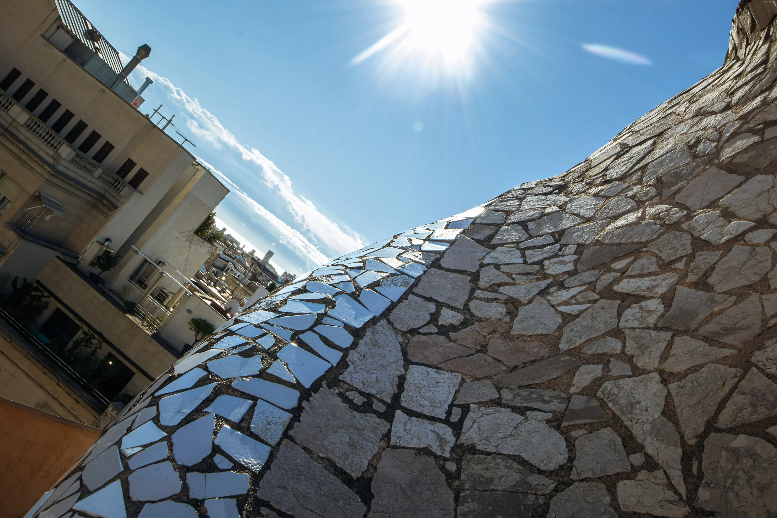 Close-up view of the mosaic-covered rooftop of Casa Milà in Barcelona, with irregular stone patterns curving toward the sky and surrounding city buildings visible in the background.
