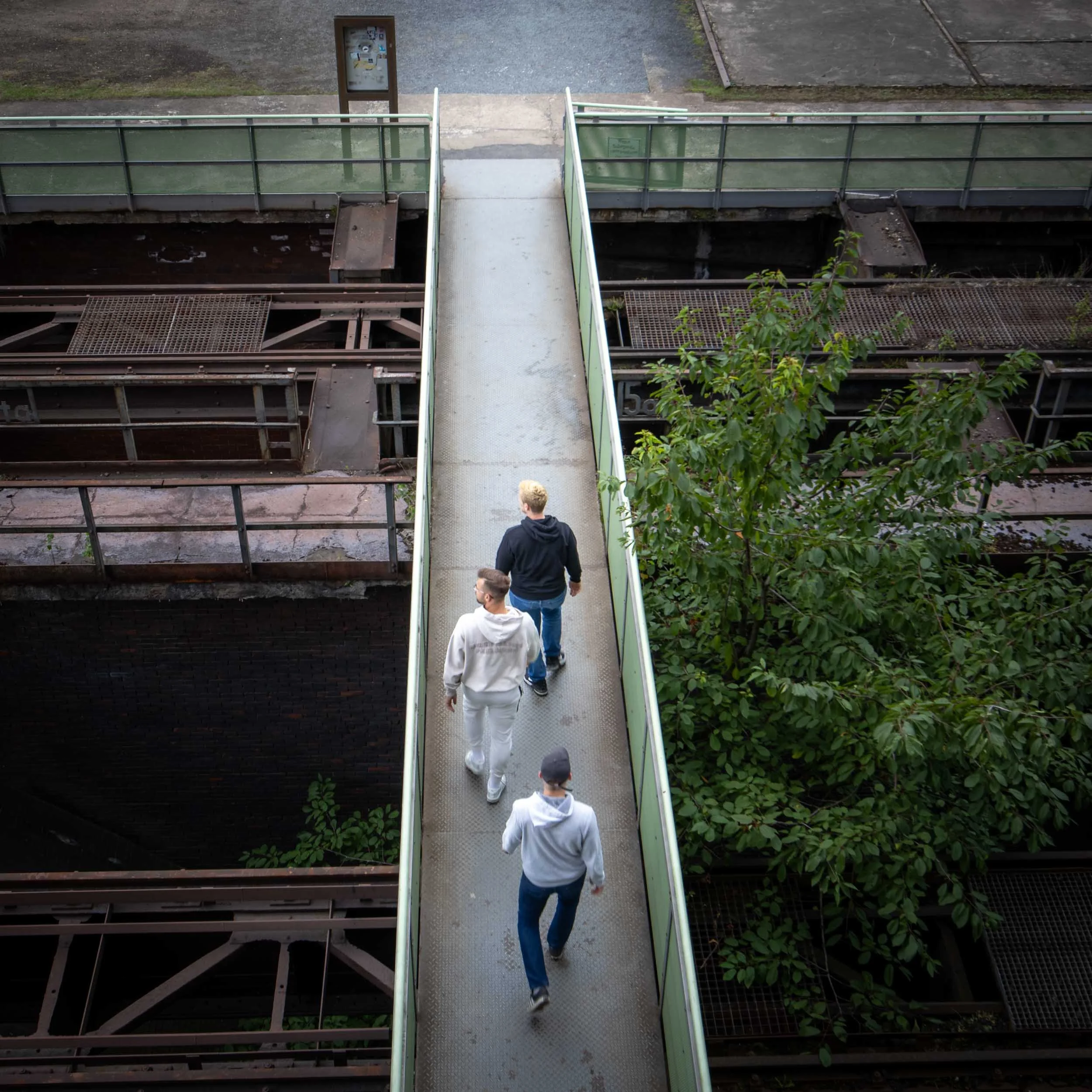 Three people walking across a narrow pedestrian bridge above an industrial structure, viewed from above.
