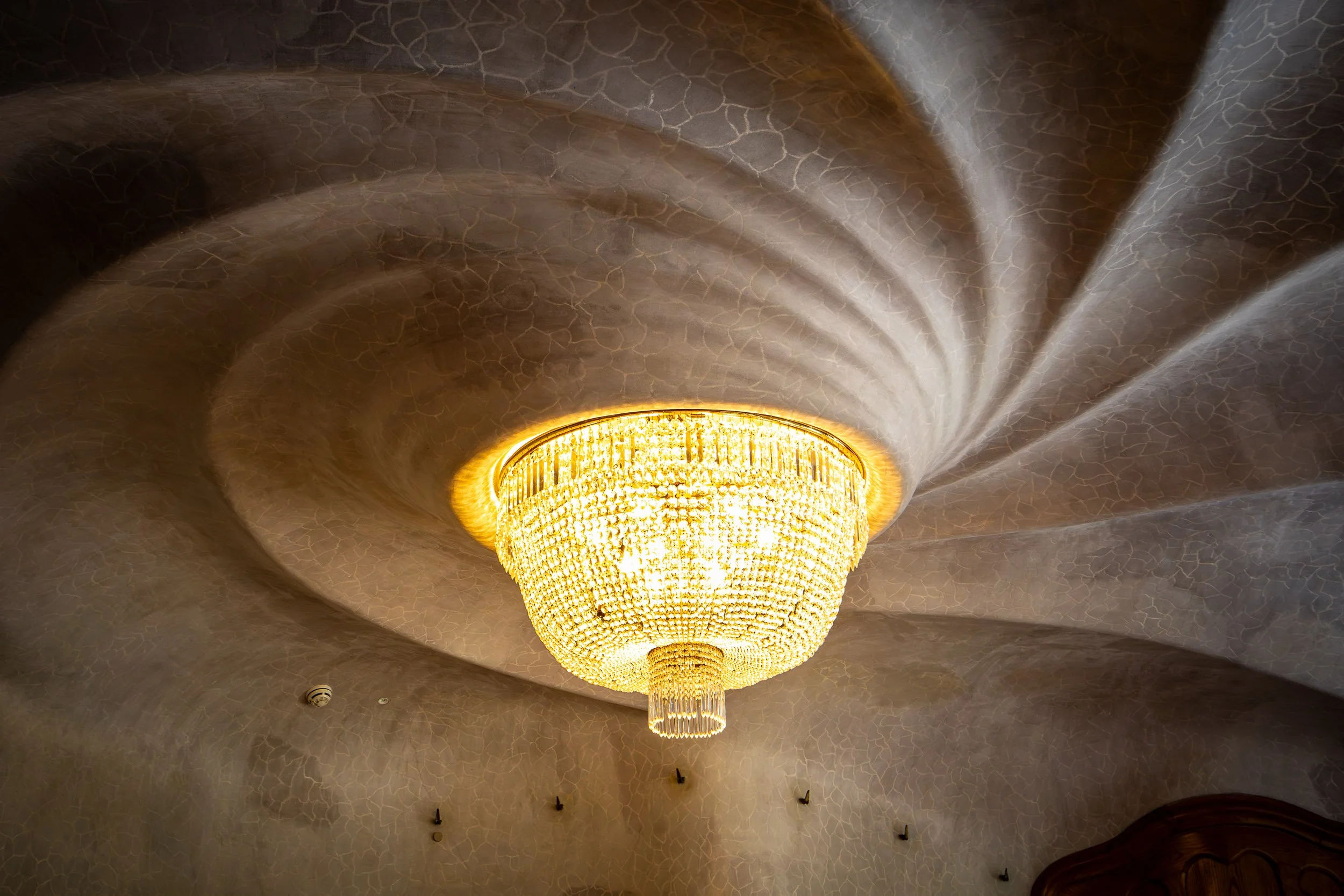 View of a crystal chandelier inside Casa Batlló in Barcelona, casting radial light patterns across a textured ceiling with organic, flowing shapes.