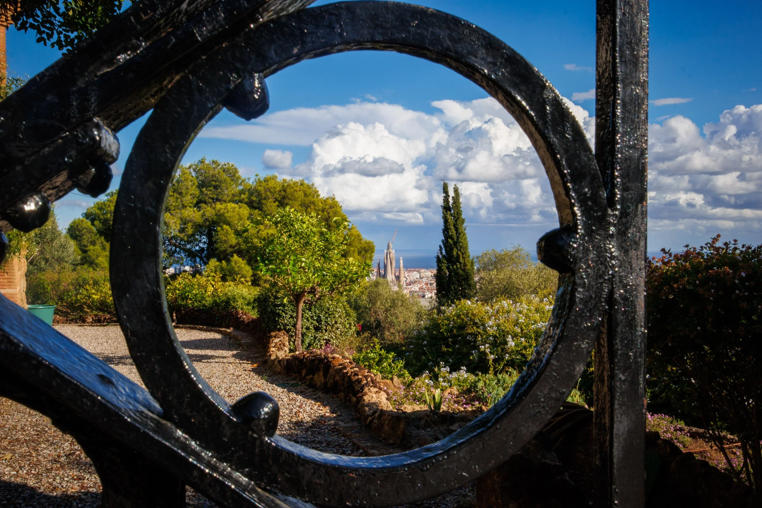 View through a circular iron railing in Parc Güell, framing greenery and a distant view of Barcelona with the towers of La Sagrada Família visible on the horizon.