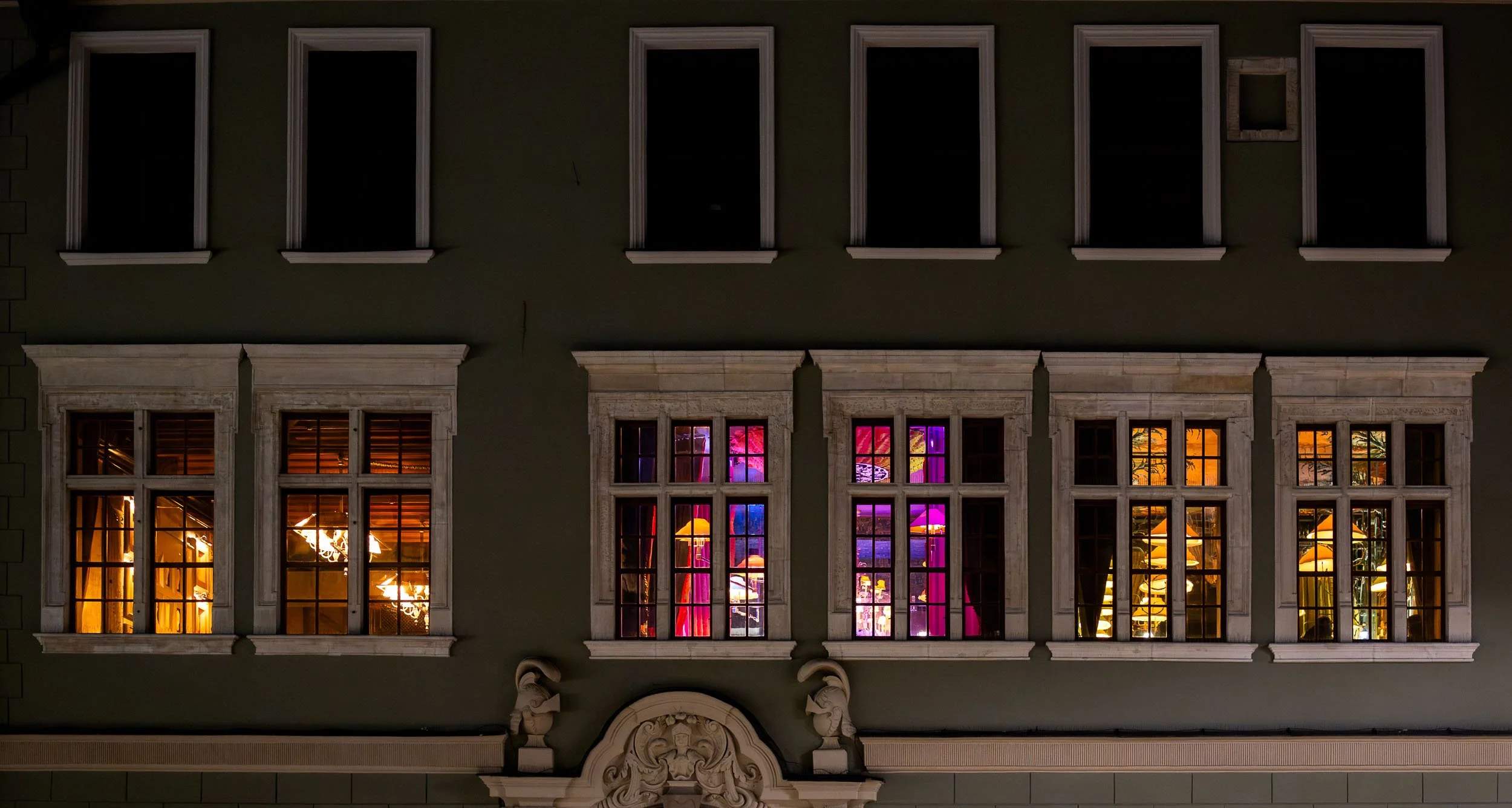 Row of illuminated windows in a historic building in Krakow at night, revealing warm and colorful interiors behind dark window frames.