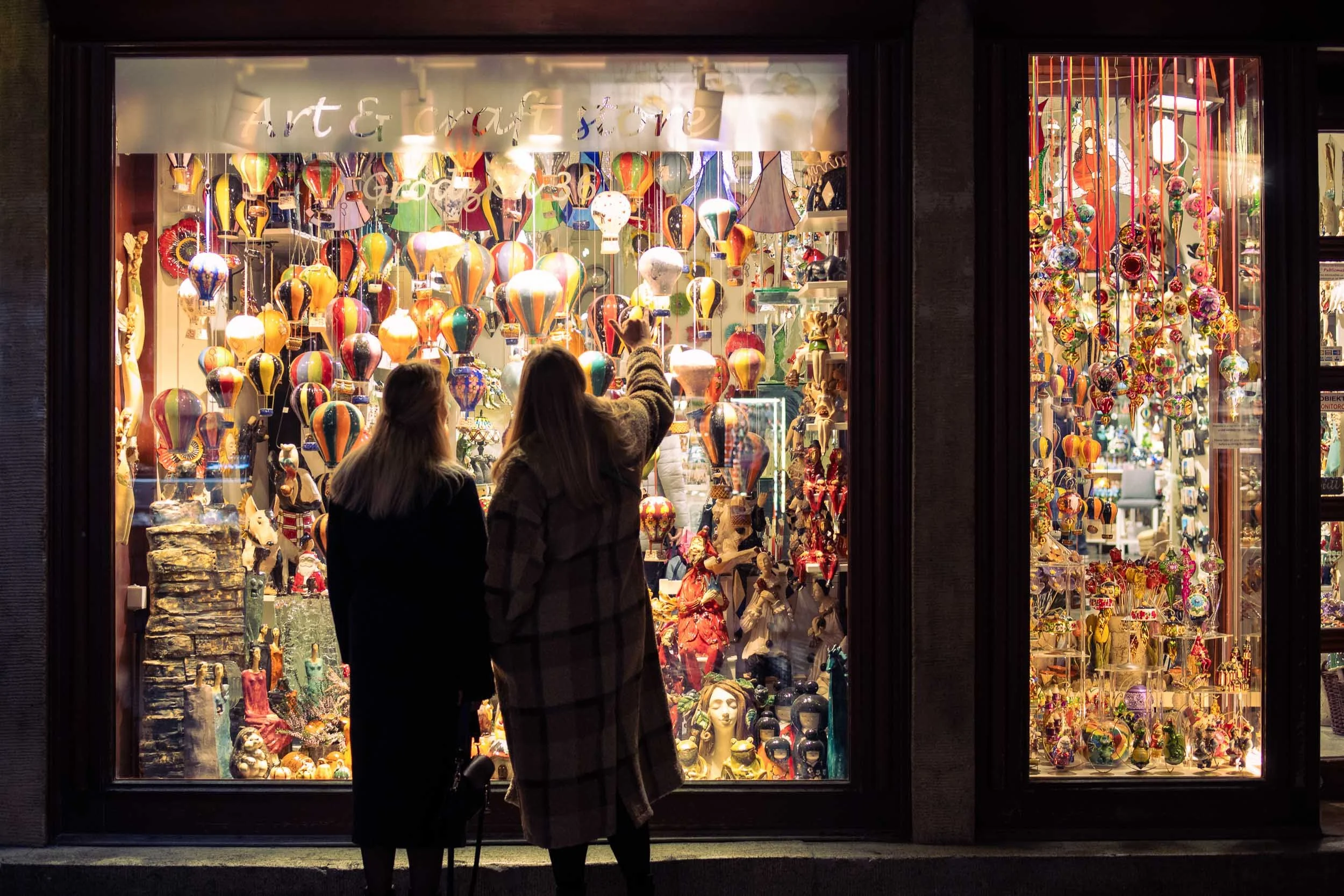 Two people standing in front of a brightly lit shop window in Krakow at night, looking at a colorful display of decorative objects and glass ornaments.