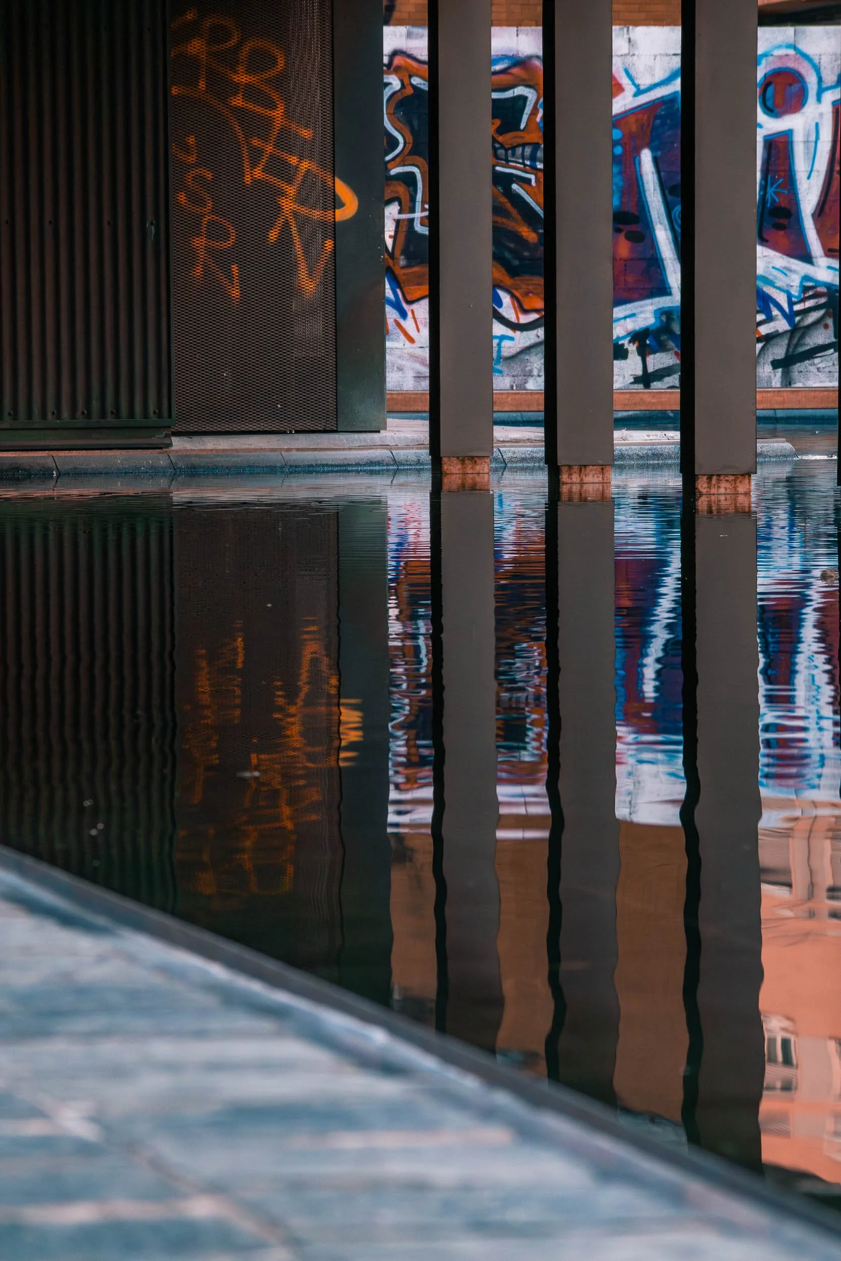 Vertical columns reflected in still water, with graffiti-covered walls in the background in the Poble-sec neighborhood of Barcelona.