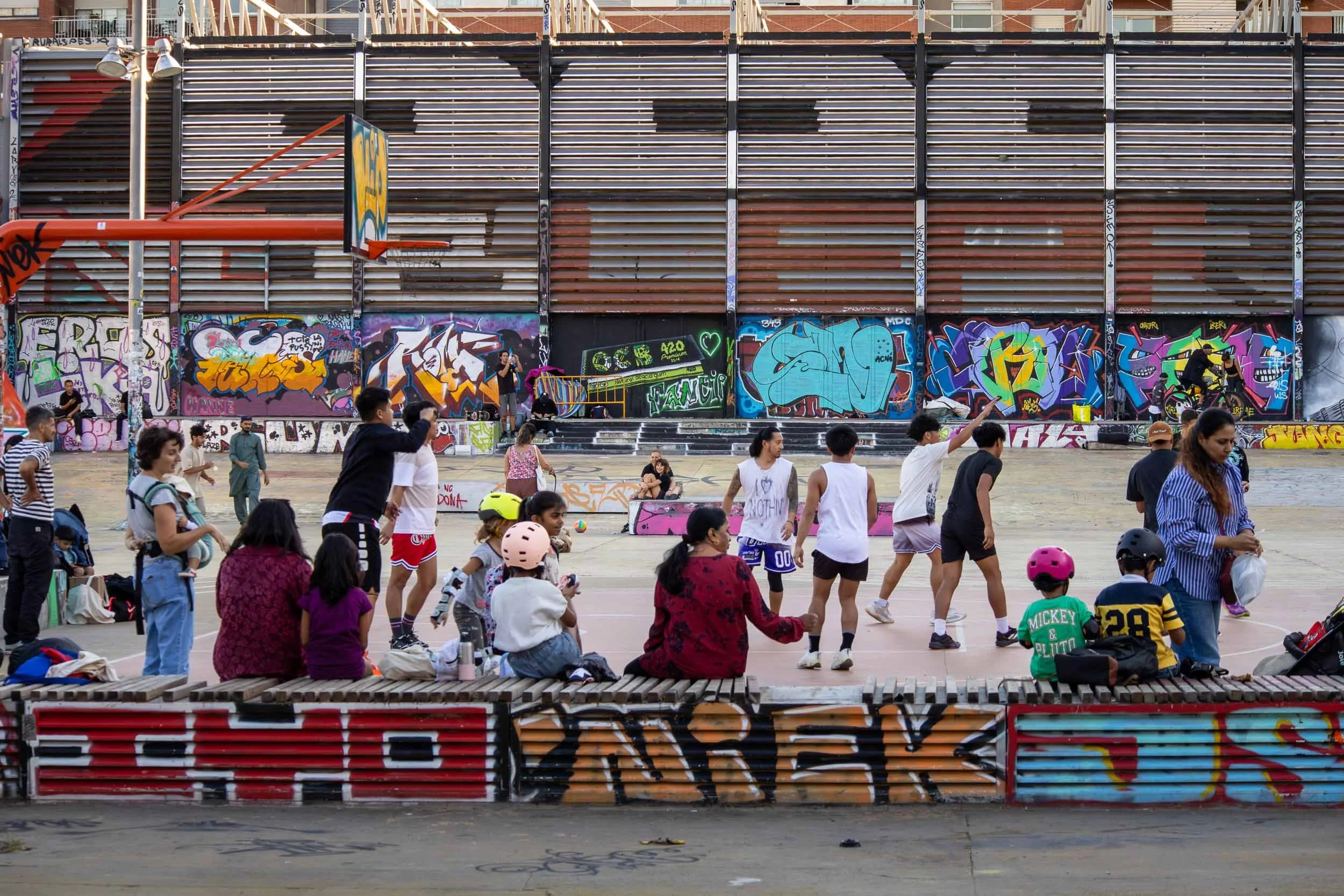 People and children gathered around an outdoor basketball court with graffiti-covered walls in the Poble-sec neighborhood of Barcelona.