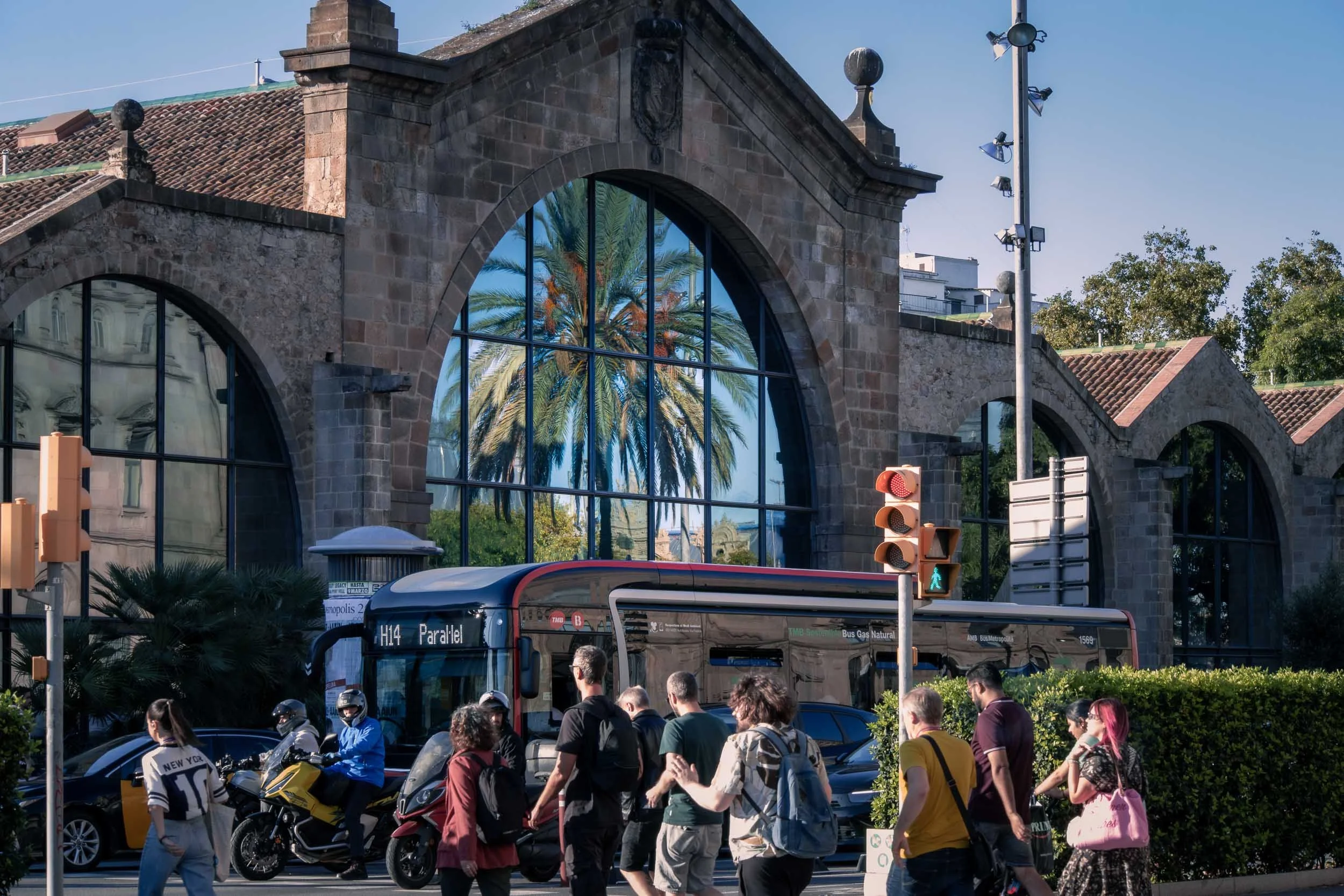 Reflection of a palm tree in the glass facade of Les Drassanes Reials, with pedestrians and a bus in the foreground.