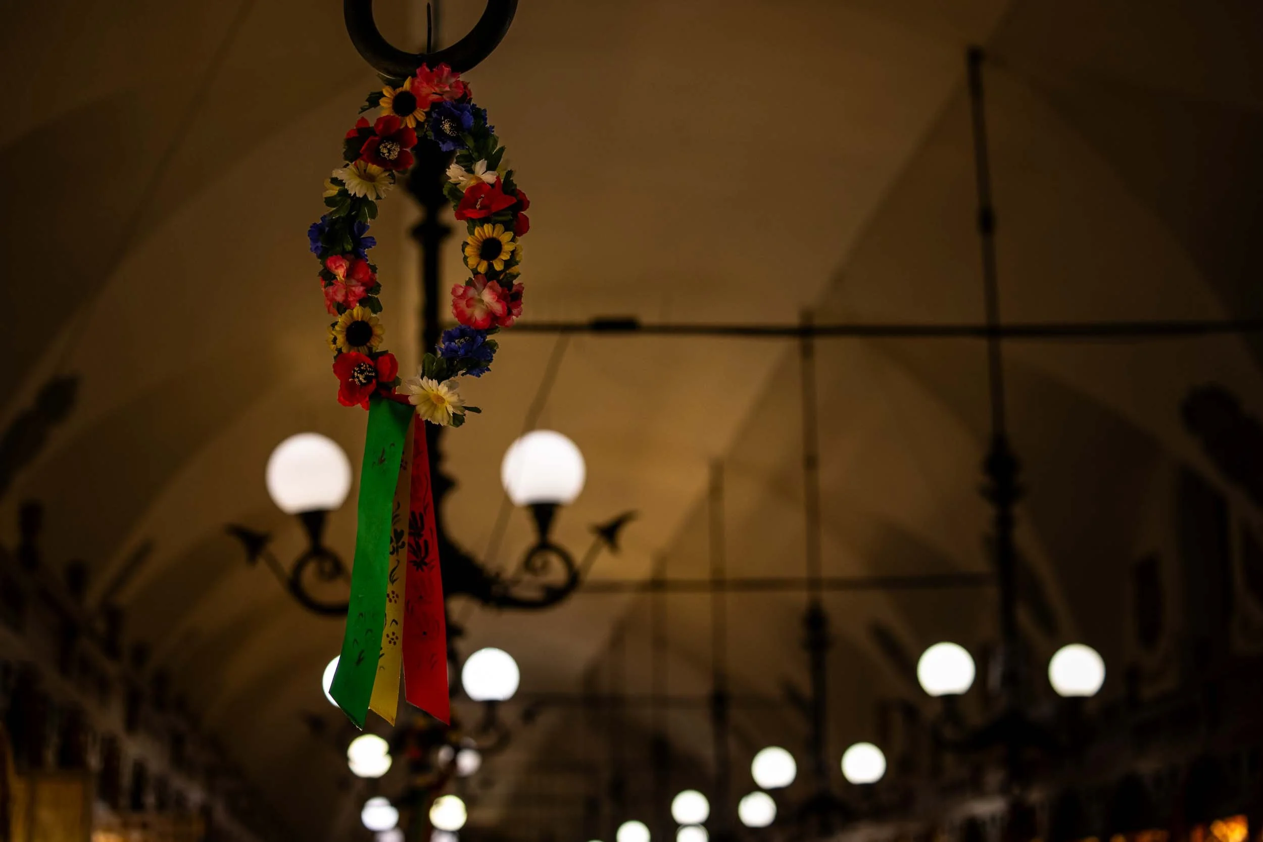 Decorative flower wreath with colorful ribbons hanging from a lamp inside the Kraków Cloth Hall, with rows of glowing lights receding into the background.
