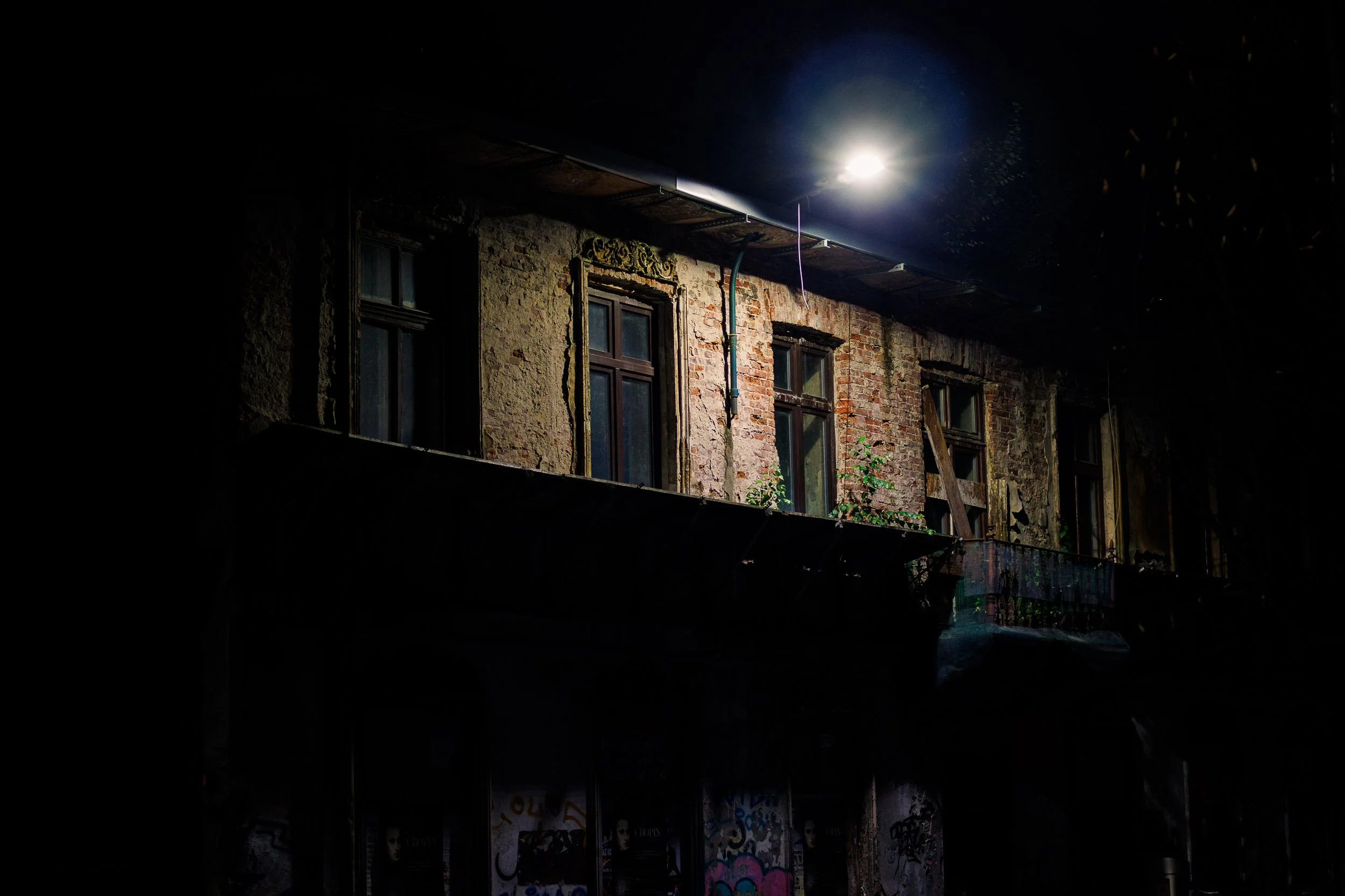 Upper floor of a weathered building in Krakow illuminated by a single streetlight at night, with worn brickwork, old windows, and graffiti visible below in the darkness.