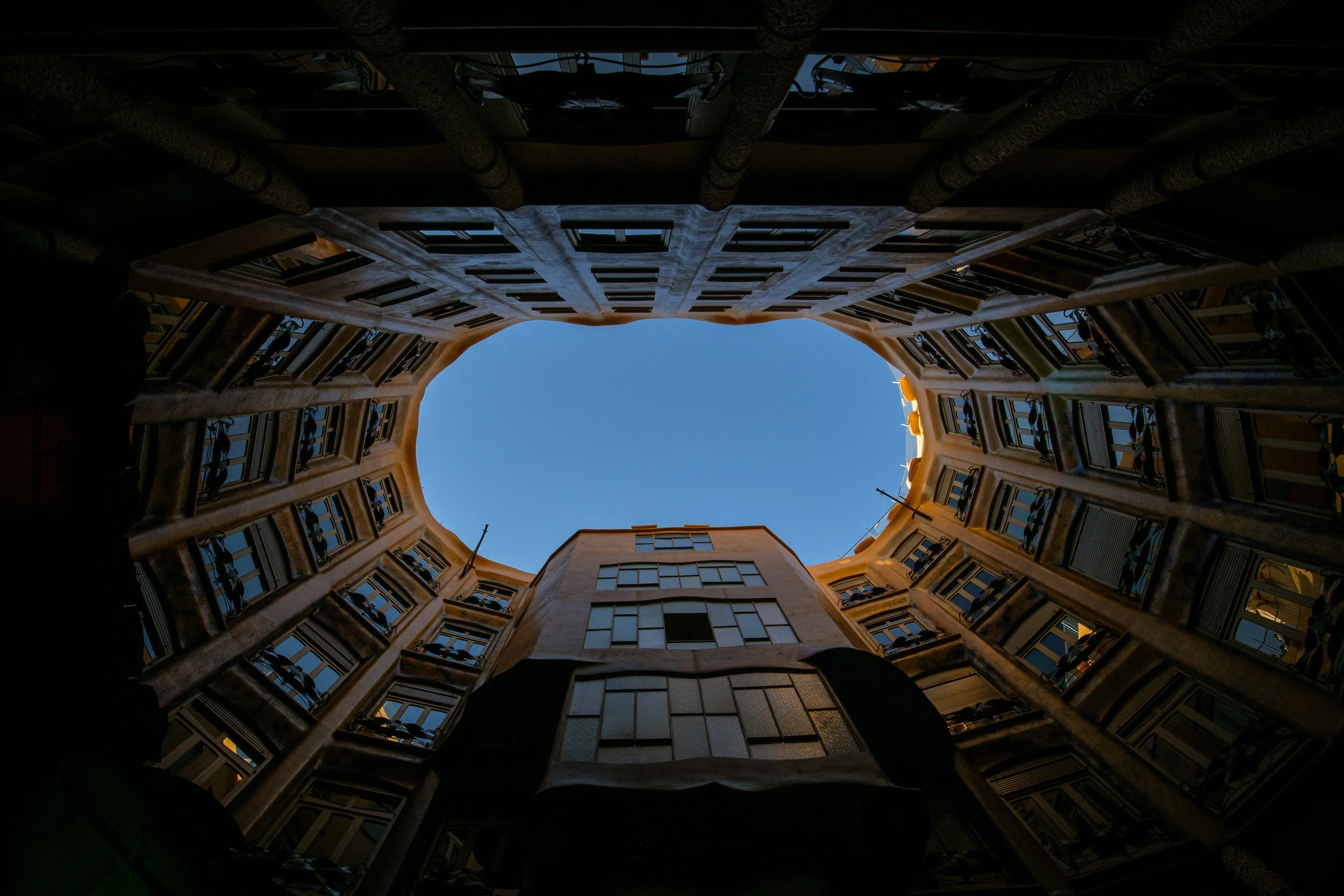 Upward view of the inner courtyard of Casa Milà in Barcelona, showing curved facades, repeating windows, and an opening to the blue sky above.