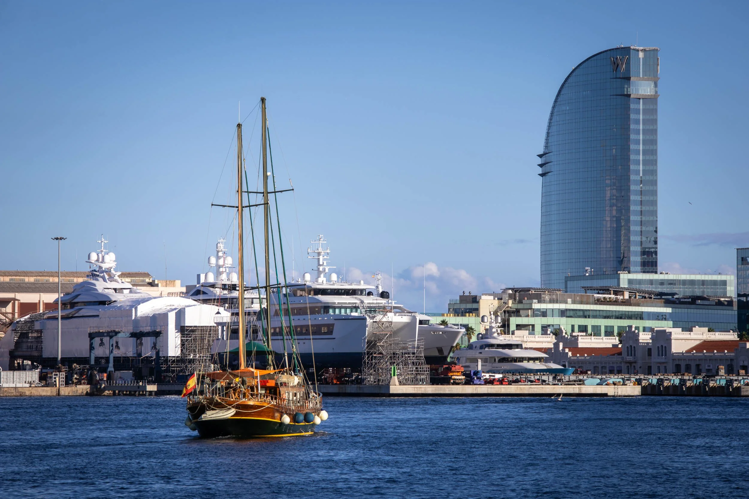 Sailboat on the water in the Port of Barcelona, with yachts and modern waterfront buildings in the background.