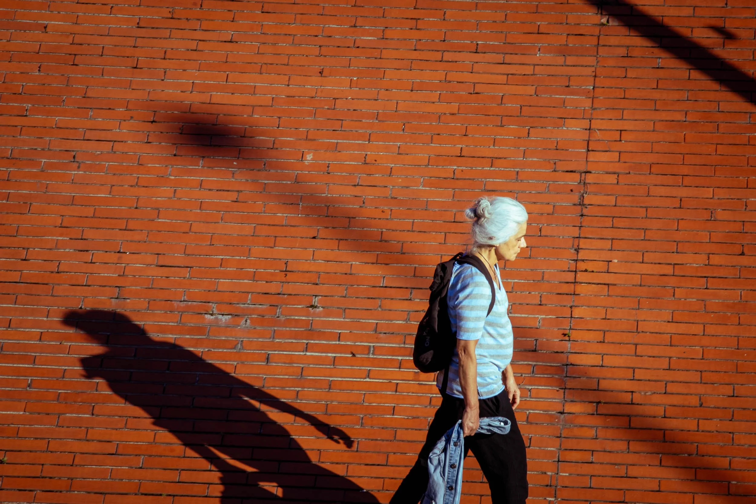 Older woman walking alongside a red brick wall, casting a long shadow.