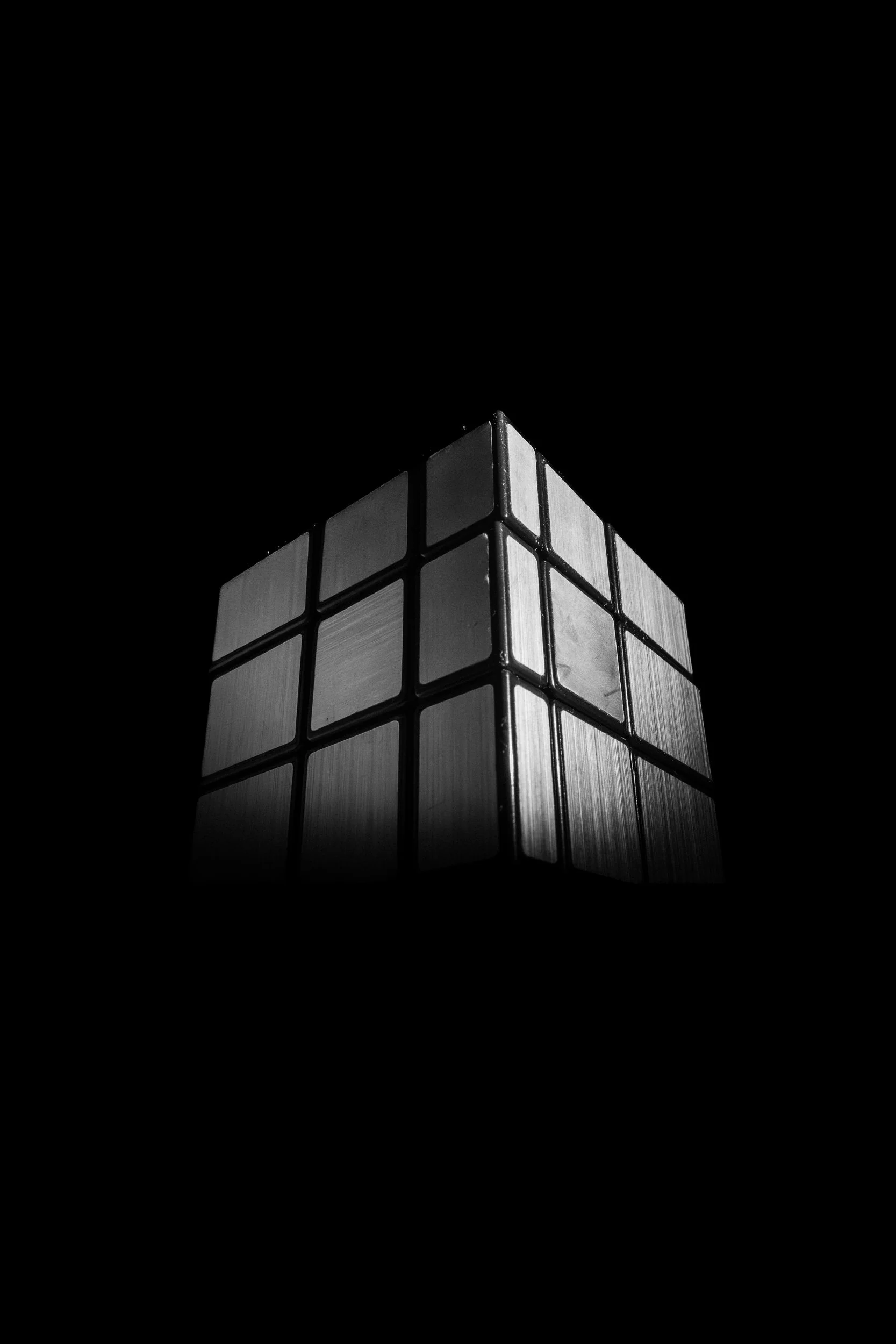 Black-and-white conceptual photograph of a Rubik's Cube emerging from darkness, illuminated by a single beam of light that emphasizes its geometric form.