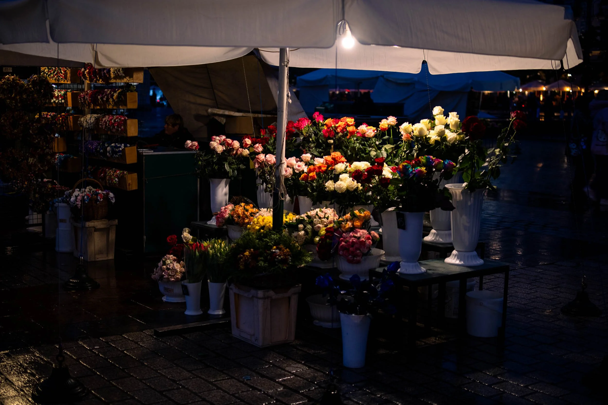 Flower stall with colorful bouquets displayed under a canopy at night in Krakow, illuminated by a single light and reflected on wet cobblestone pavement.