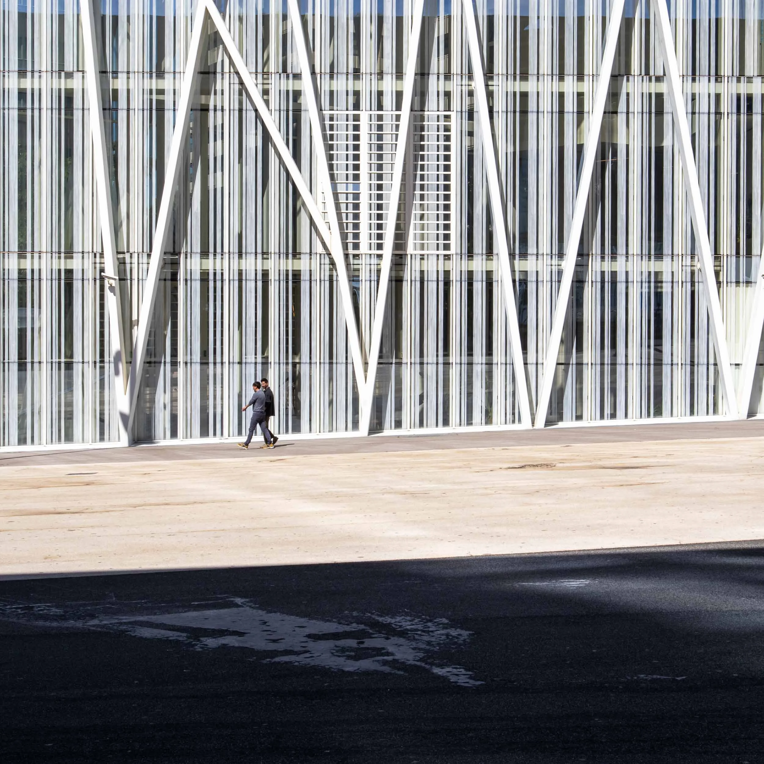 Two people walking in front of a modern building with a glass facade and white structural beams.