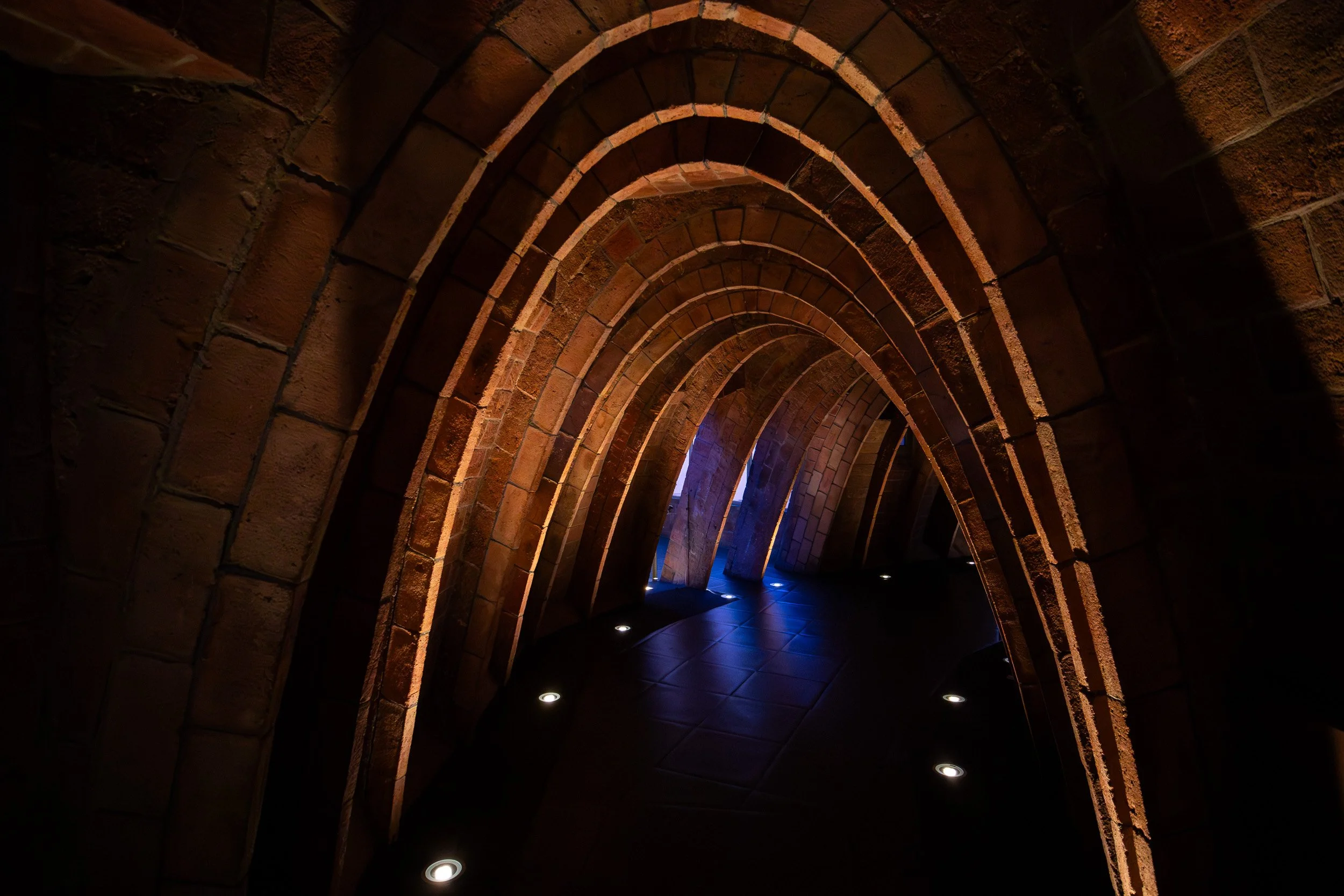 Series of stone arches inside Casa Milà in Barcelona, forming a curved passage with warm lighting and a tiled floor illuminated by small recessed lights.