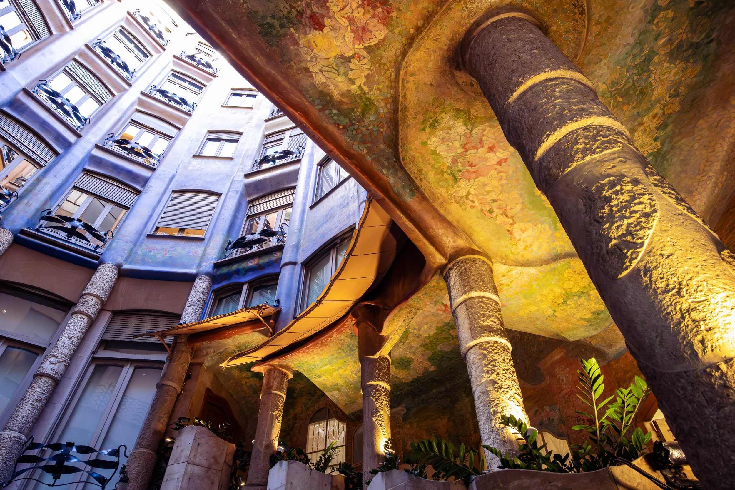 Interior view of Casa Milà in Barcelona showing textured stone columns, organic ceiling patterns, and curved facades illuminated by warm light from below.