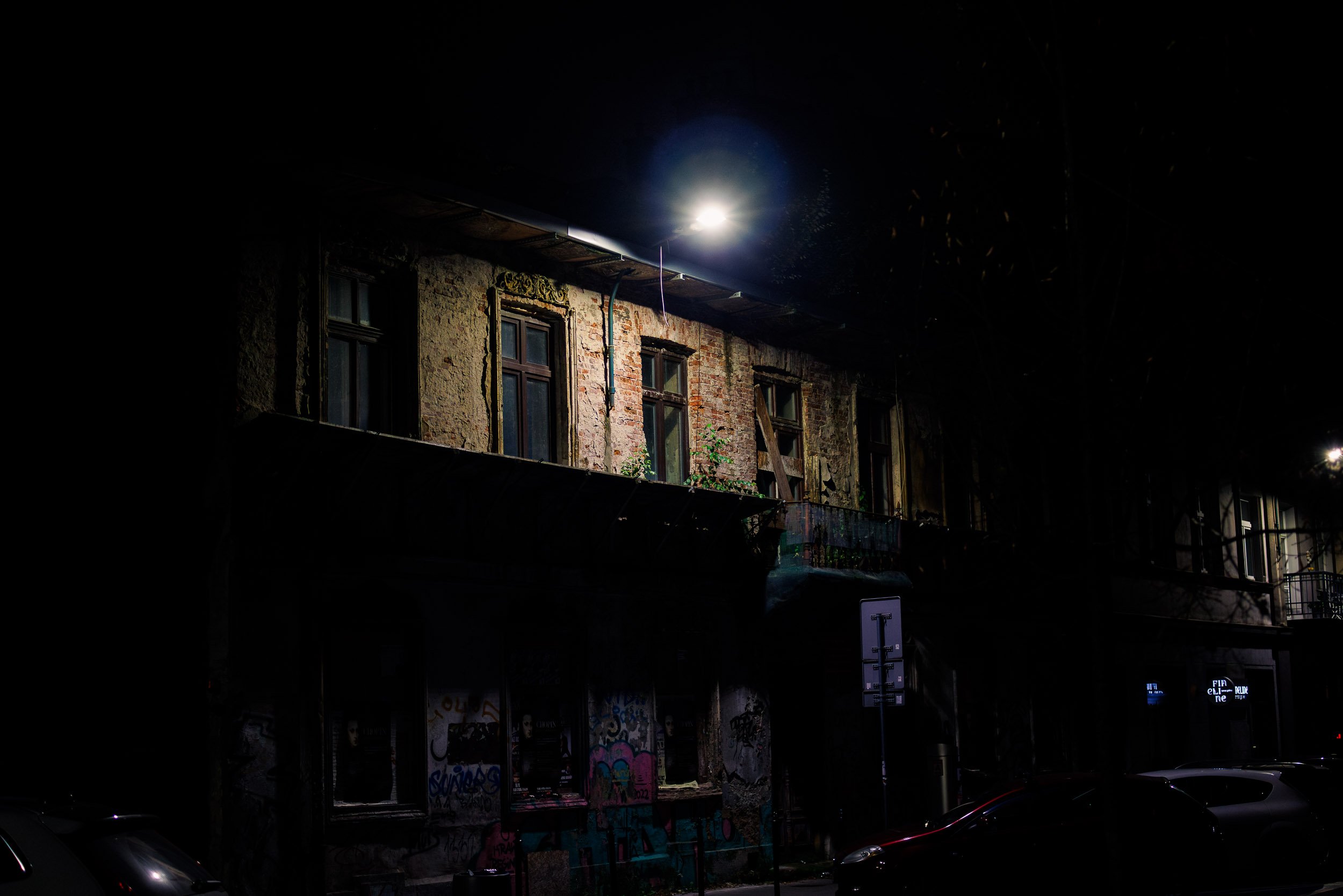 Upper floor of a weathered building in Krakow illuminated by a single streetlight at night, with worn brickwork, old windows, and graffiti visible below in the darkness.