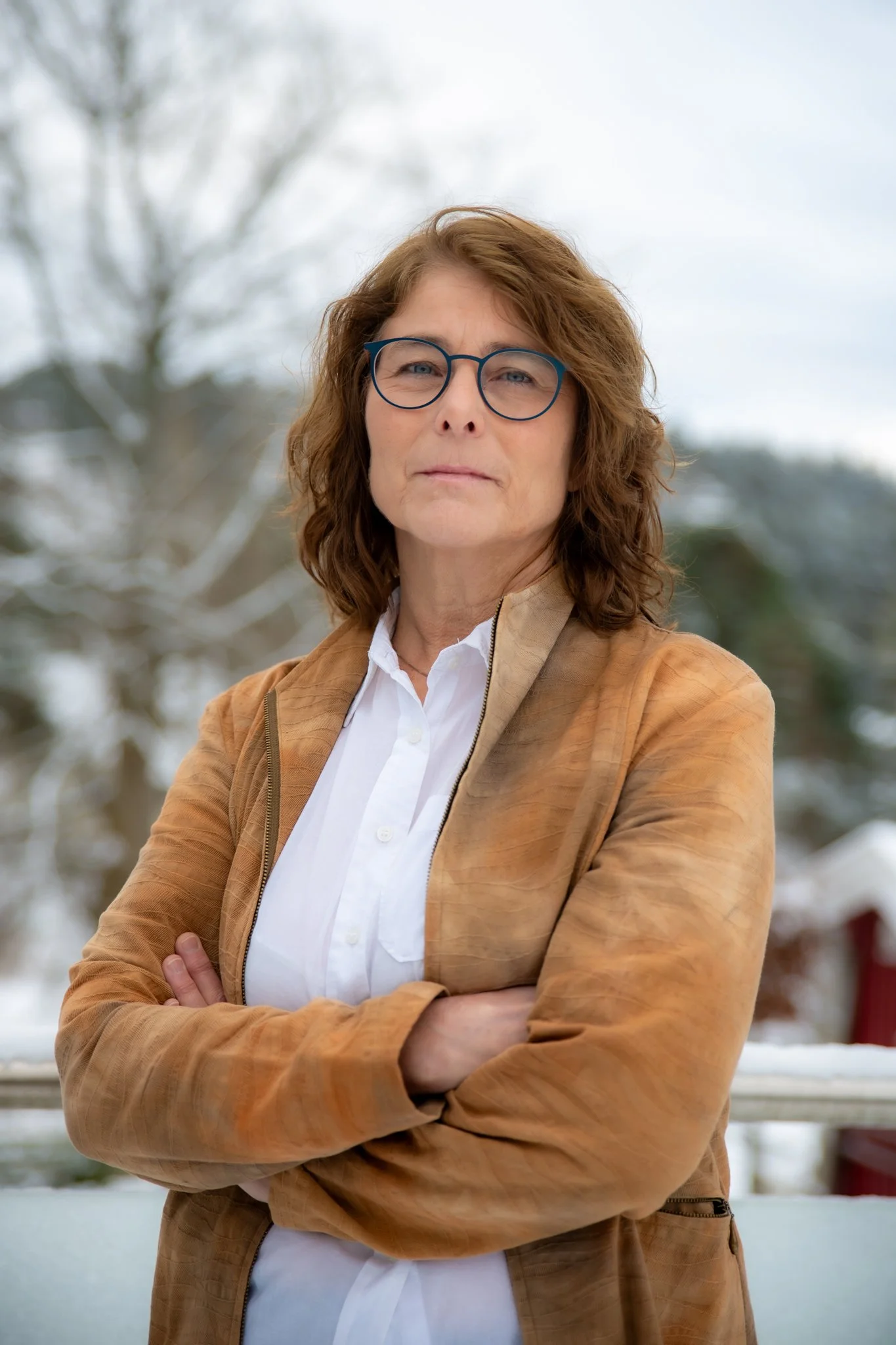 Kvinne med brunt, løst hår, iført hvit skjorte og brunt skinnjakke, står med armene overkors utendørs med snødekte trær i bakgrunnen.